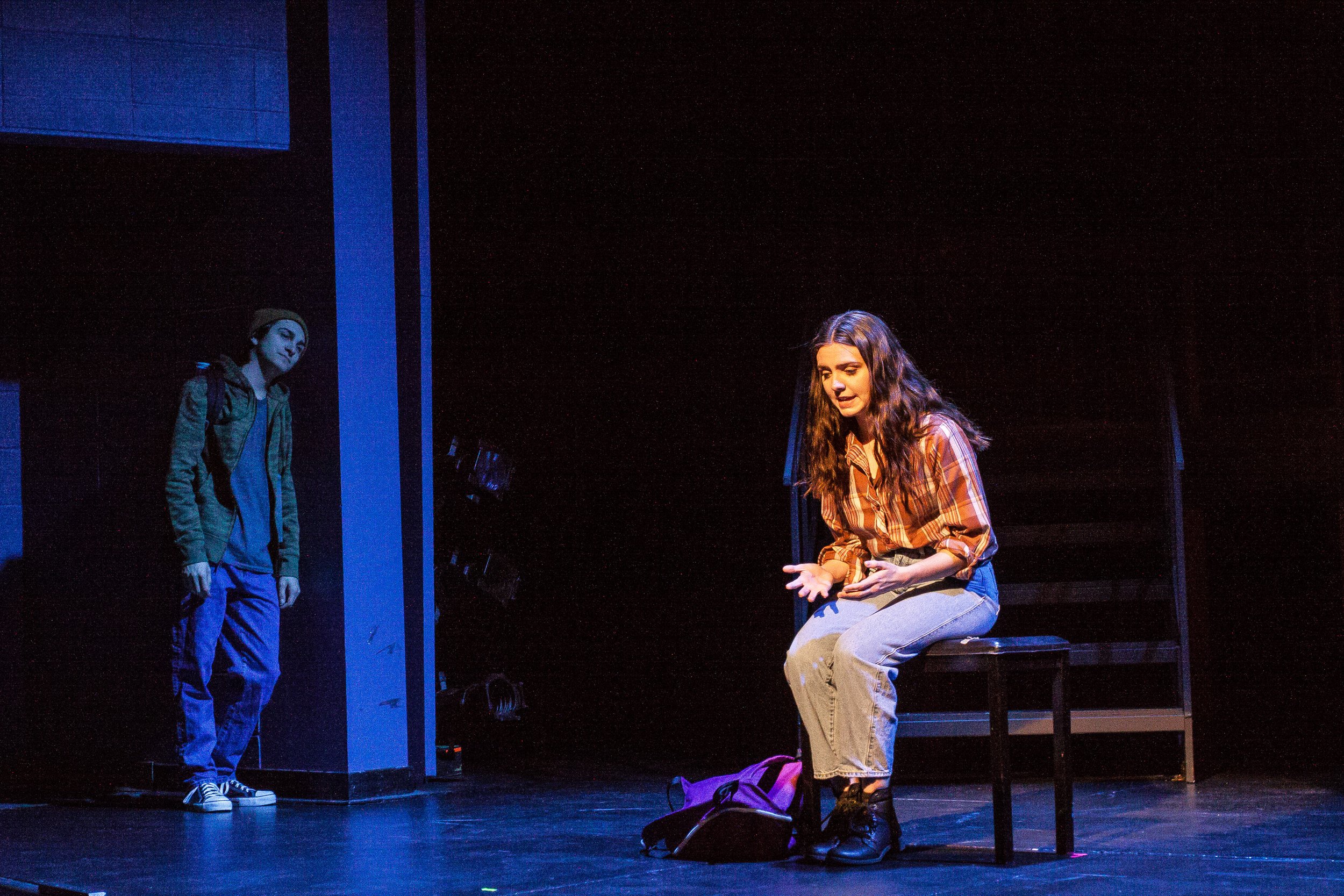 Two young women are performing on stage, one sitting on a chair and speaking, the other standing in the background, listening.