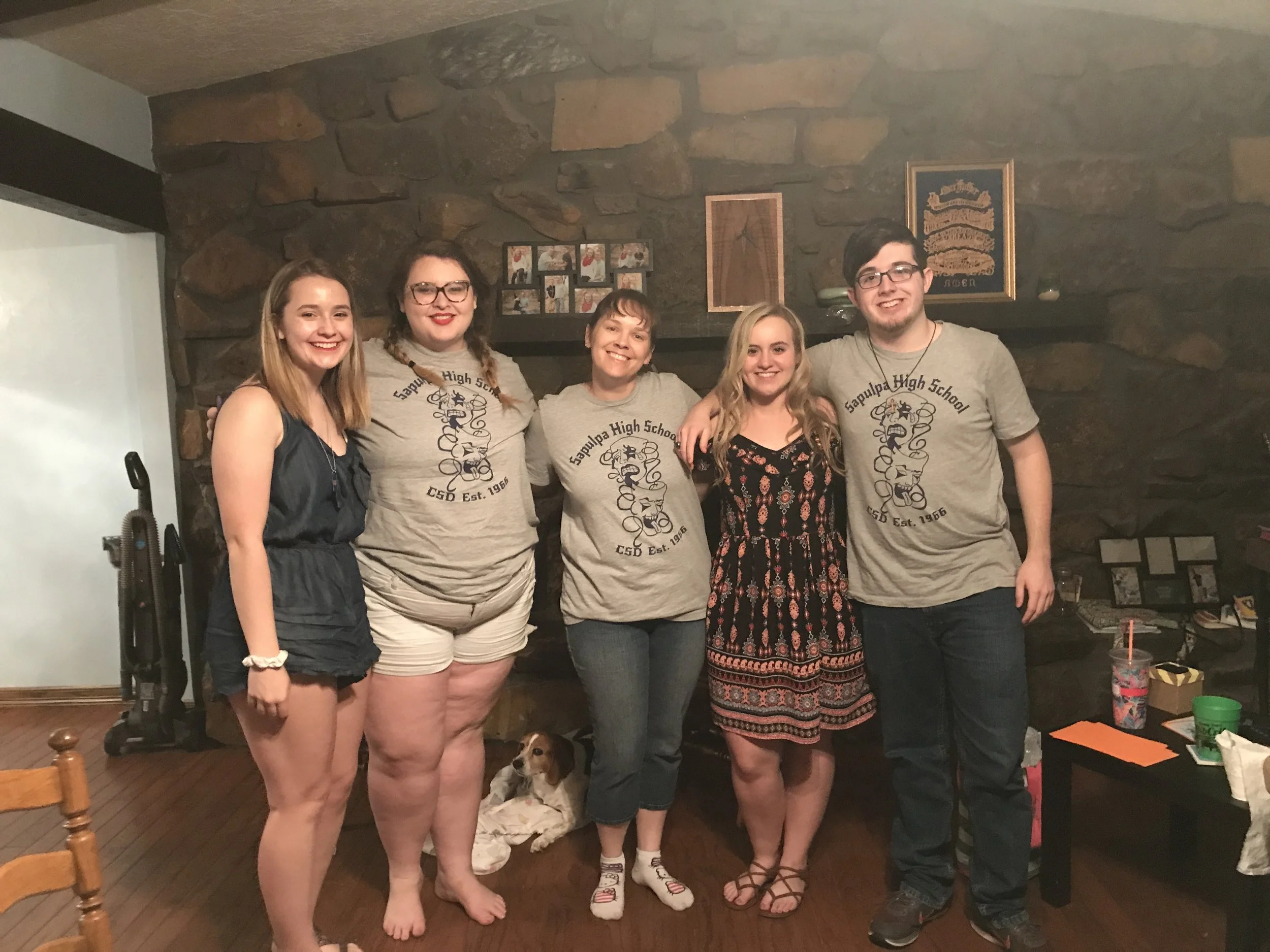 Five young adults standing together in a living room, smiling at the camera. Behind them is a stone fireplace with framed photos and decorations. A small dog is lying on the floor near them. The group appears to be celebrating or gathering casually.