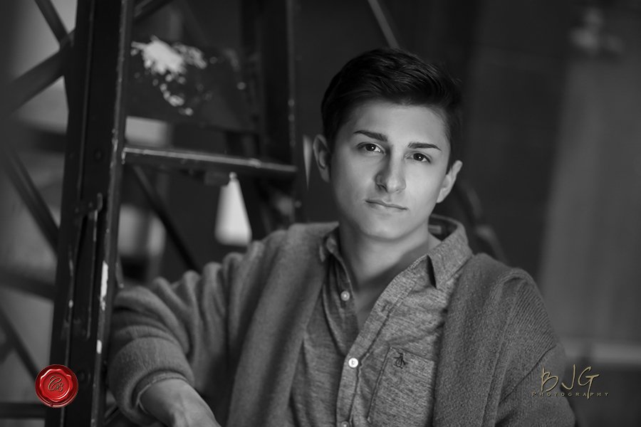 Black and white portrait of a young man with styled dark hair, wearing a collared shirt and cardigan, leaning against a metal structure.