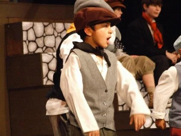 Children dressed in costumes performing in a play, with a boy in the foreground wearing a vest, shirt, scarf, and cap.