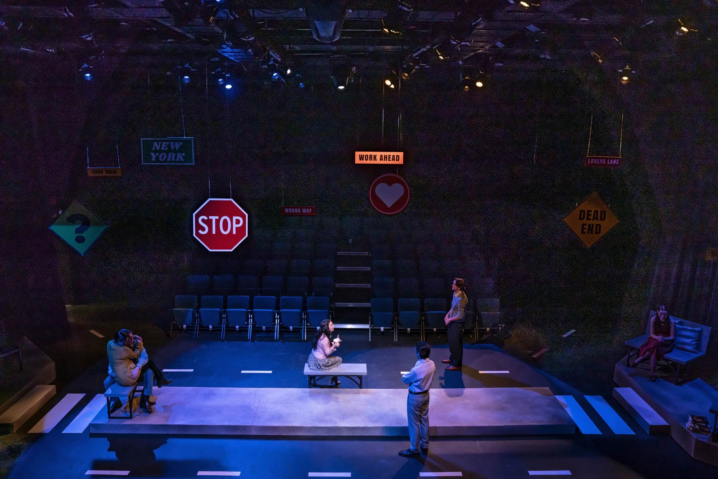 A theatrical stage with dark walls decorated with traffic signs and signals, including a stop sign, a question mark, and signs indicating 'Wrong Way,' 'Work Ahead,' 'Lovers Lane,' and 'Dead End.' There are five people on stage, some seated and others