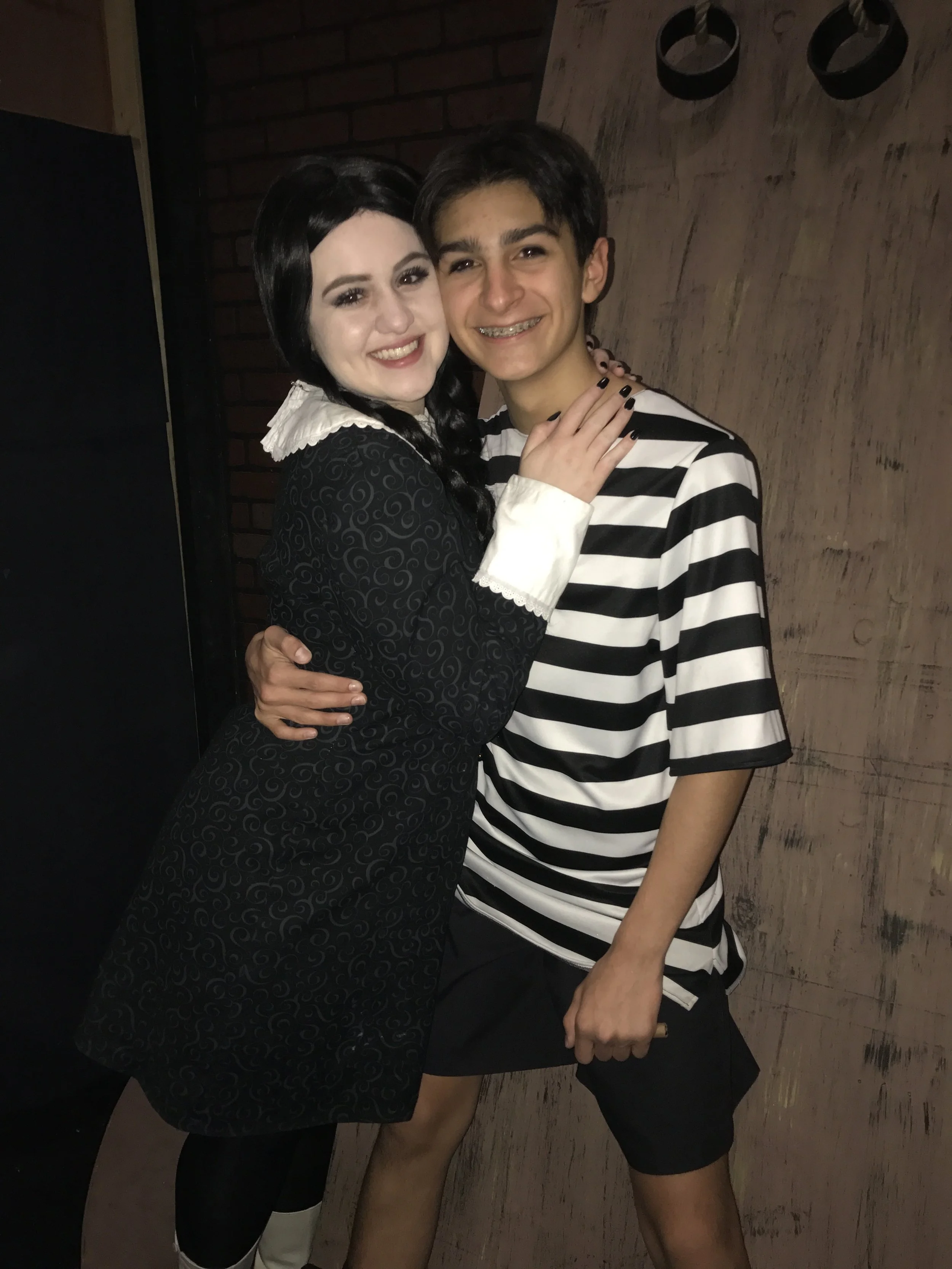 Two young people, a girl and a boy, are hugging and smiling at the camera. The girl has dark hair and is wearing a black dress with a white collar and cuffs. The boy has dark hair, braces, and is wearing a black and white striped shirt with black sho