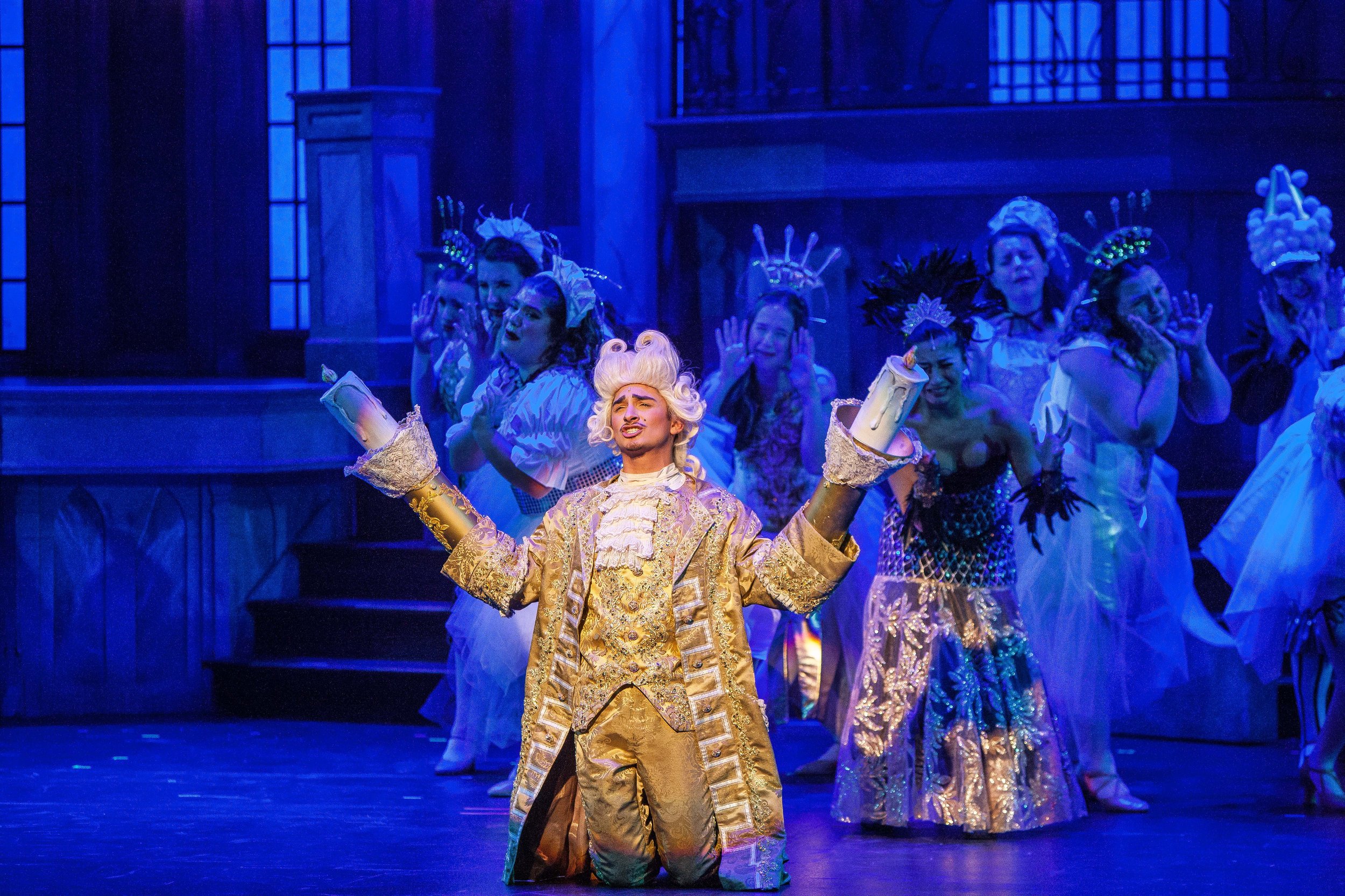 Theatrical stage scene with a performer dressed as Louis XV in a gold costume, surrounded by cast members in period costumes, under blue stage lighting.