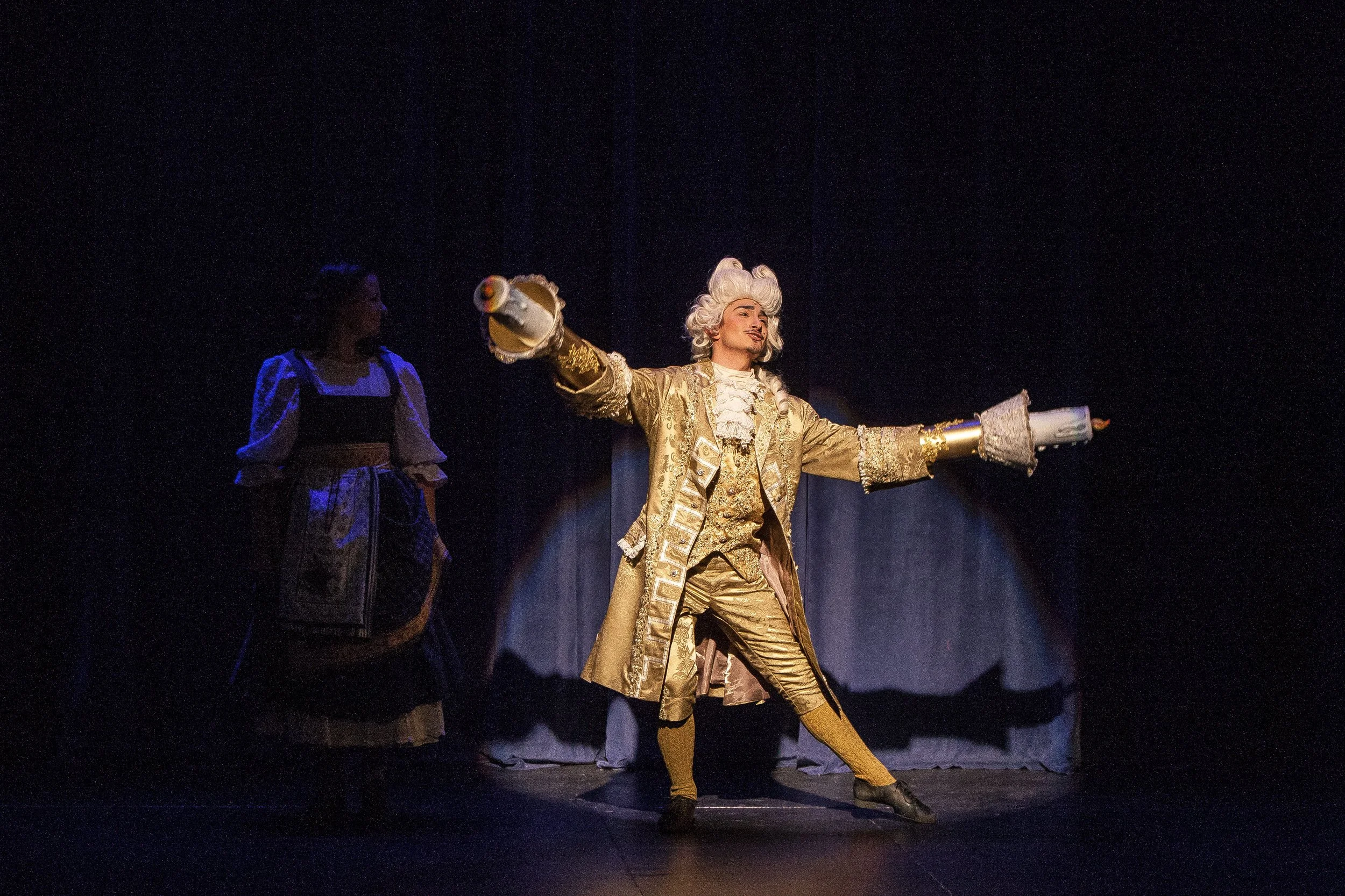 Theater scene with two actors in historical costumes; one actor in a gold brocade coat and wig holding two costumes, another in a traditional dress watching.