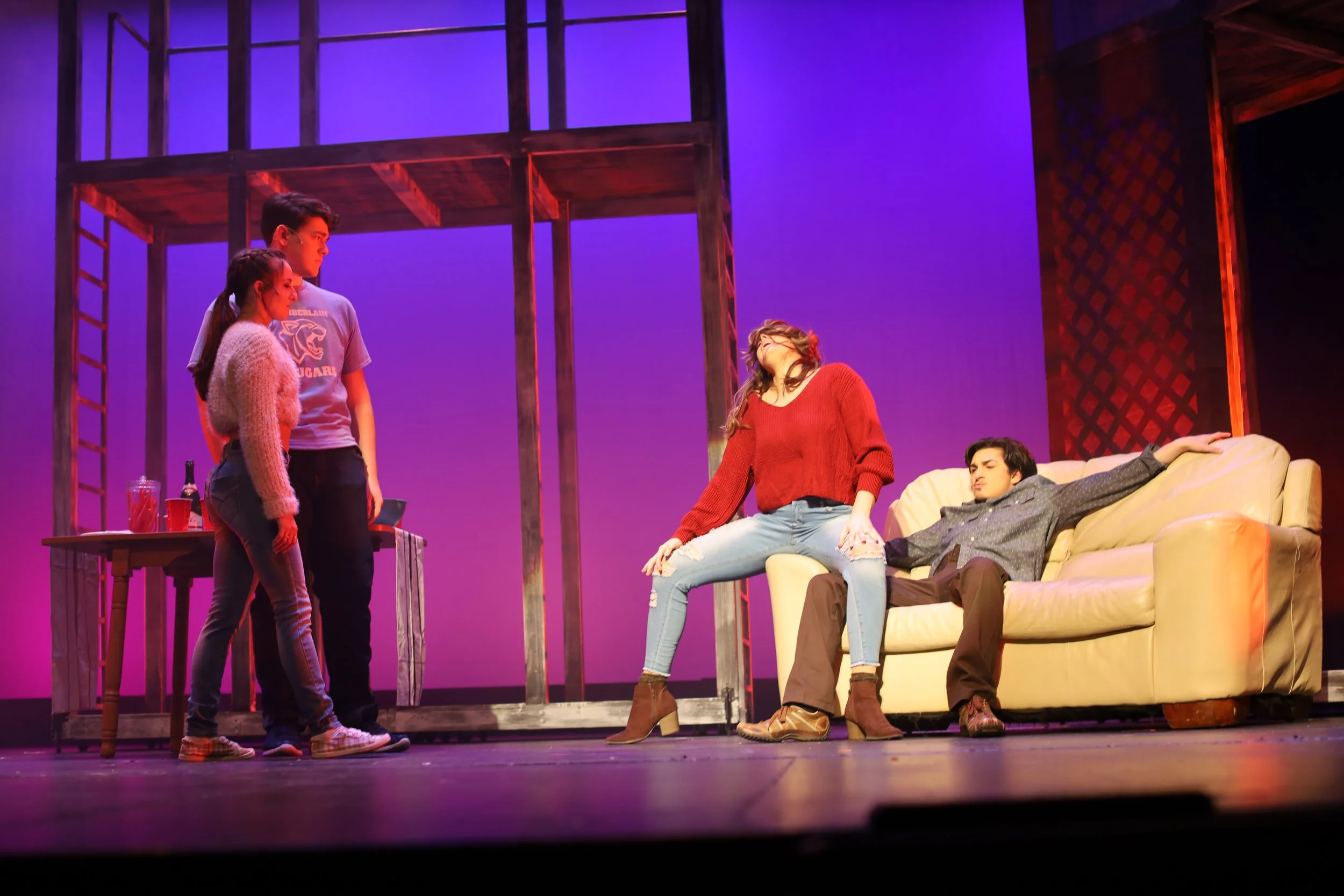Theater scene with four actors on stage, two standing by a table, one sitting on a couch, and one kneeling on the couch, in front of a purple illuminated set.
