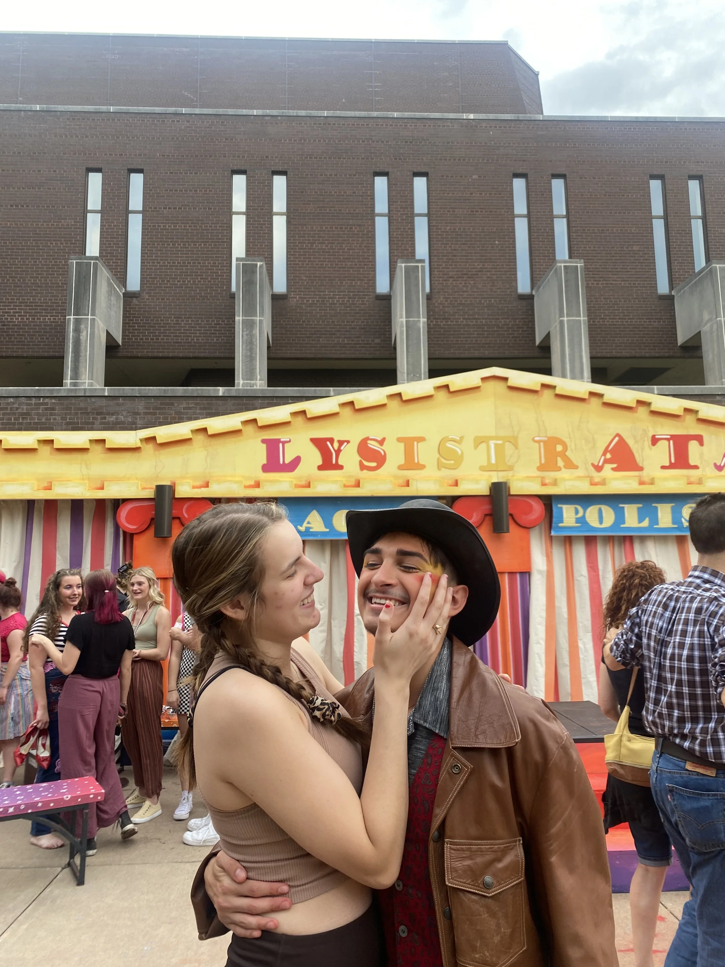 A young woman and young man are at an outdoor fair. The woman is smiling and touching the man's face, and the man is smiling with his eyes closed. They are standing in front of a colorful game booth with the word 'Lysistrata' on it, and other people 