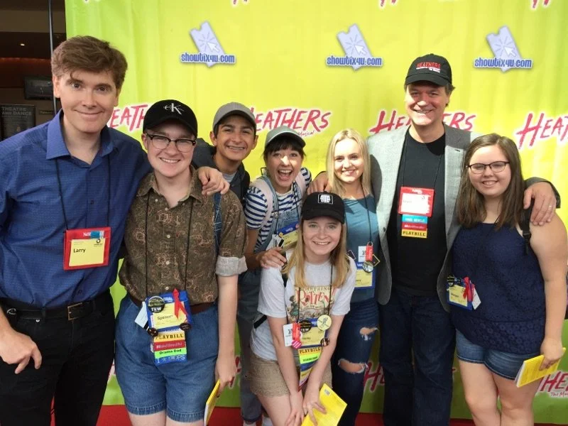 Group of seven smiling young people and two adult men standing together posed for a photo at an event with a yellow background displaying the words 'Heathers' and 'showtix4u.com'.