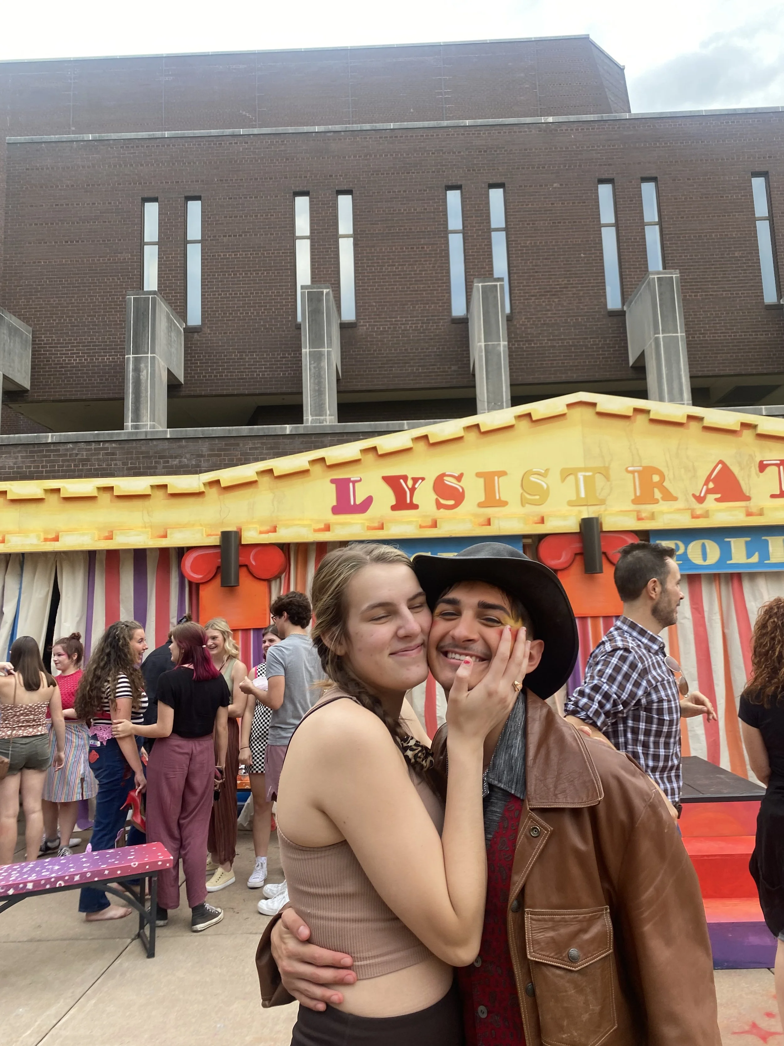 Two people hugging and smiling at a fairground game booth called 'Lighthouse'. The woman has her eyes closed and the man is wearing a hat and a leather jacket, and they are surrounded by other people.