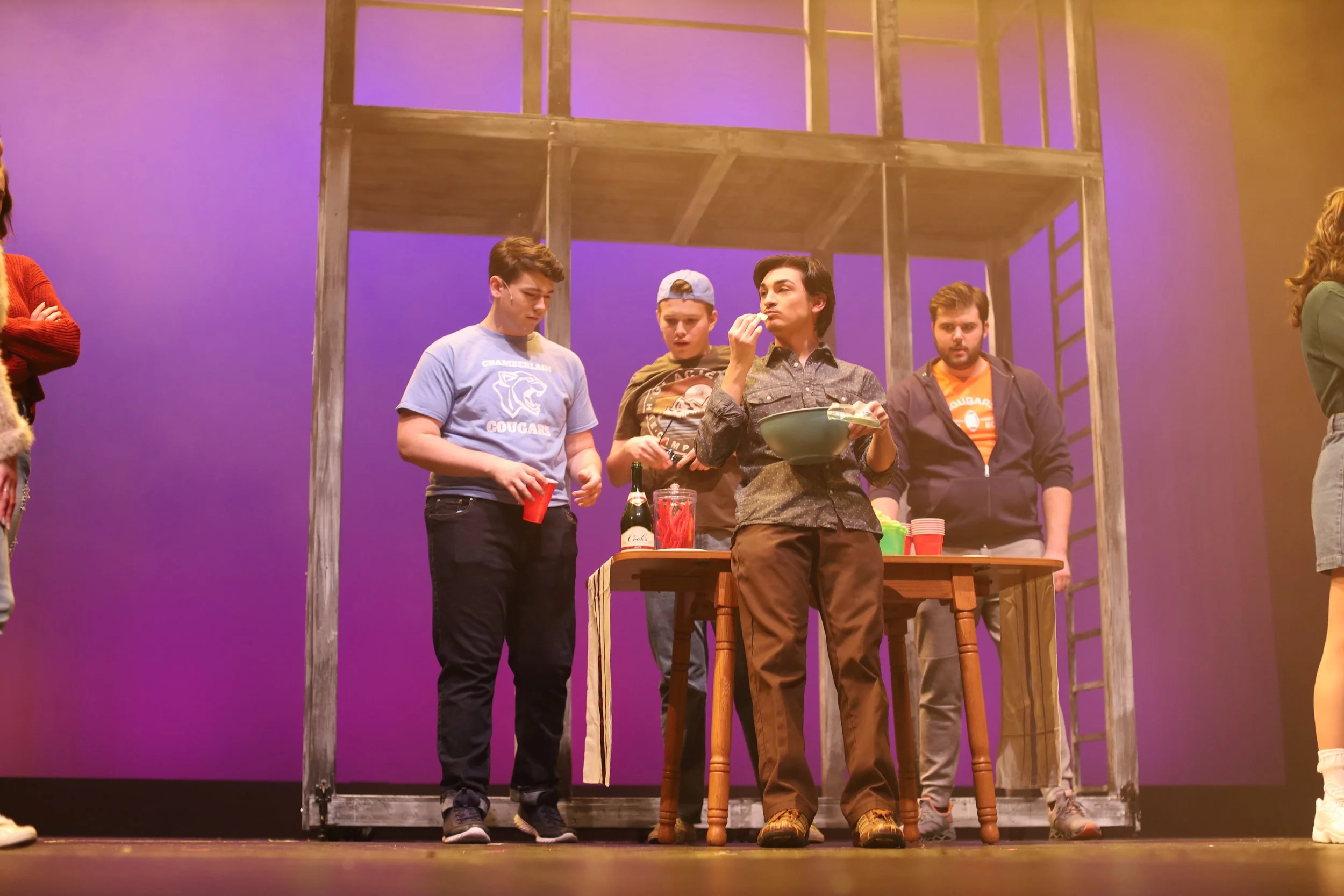 A group of young people standing near a wooden table on a stage with a purple backdrop, some are holding cups, and one person is eating from a bowl.