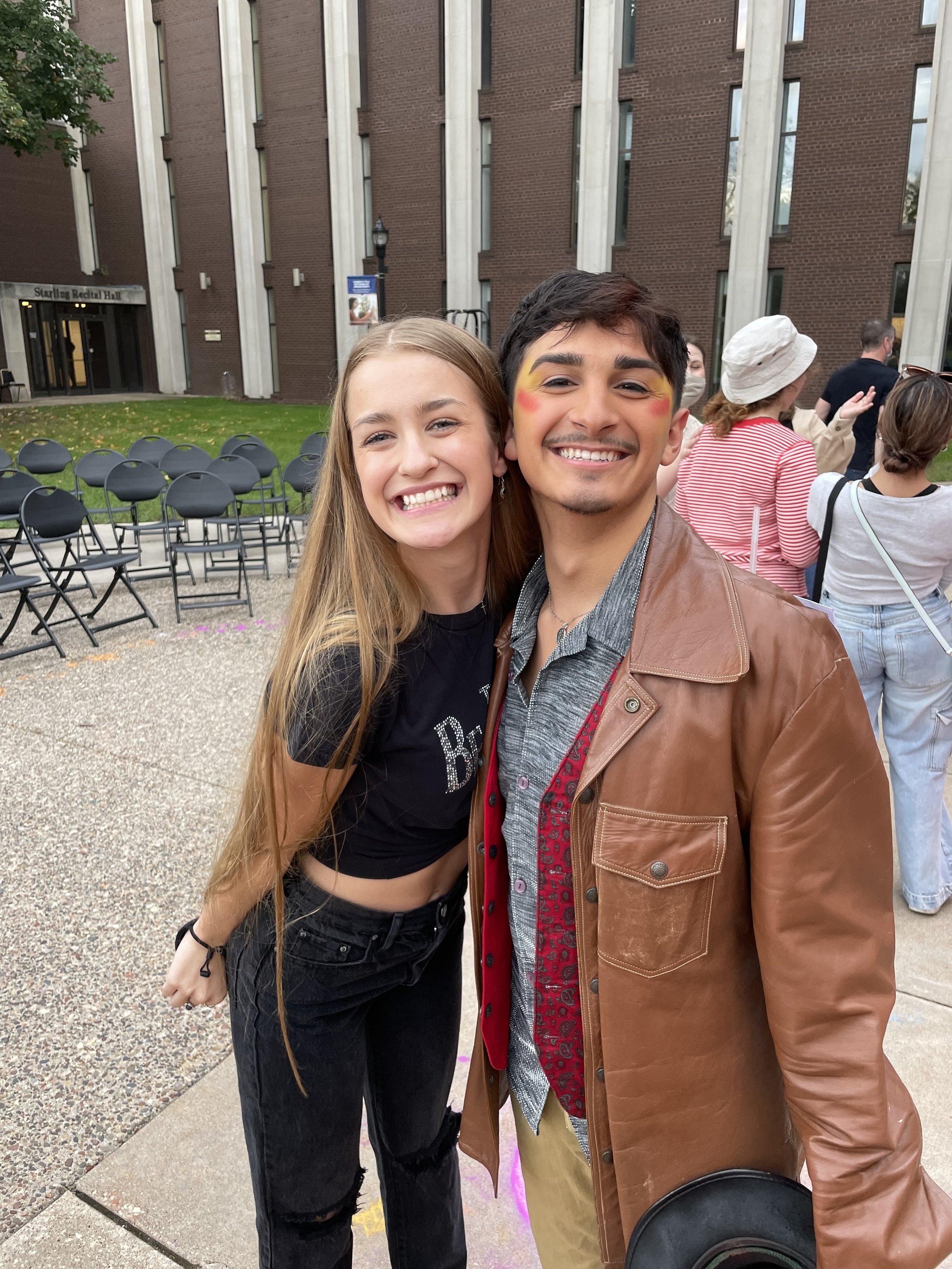 Two young people smiling and posing for a selfie outdoors, with a brick building and other people in the background.