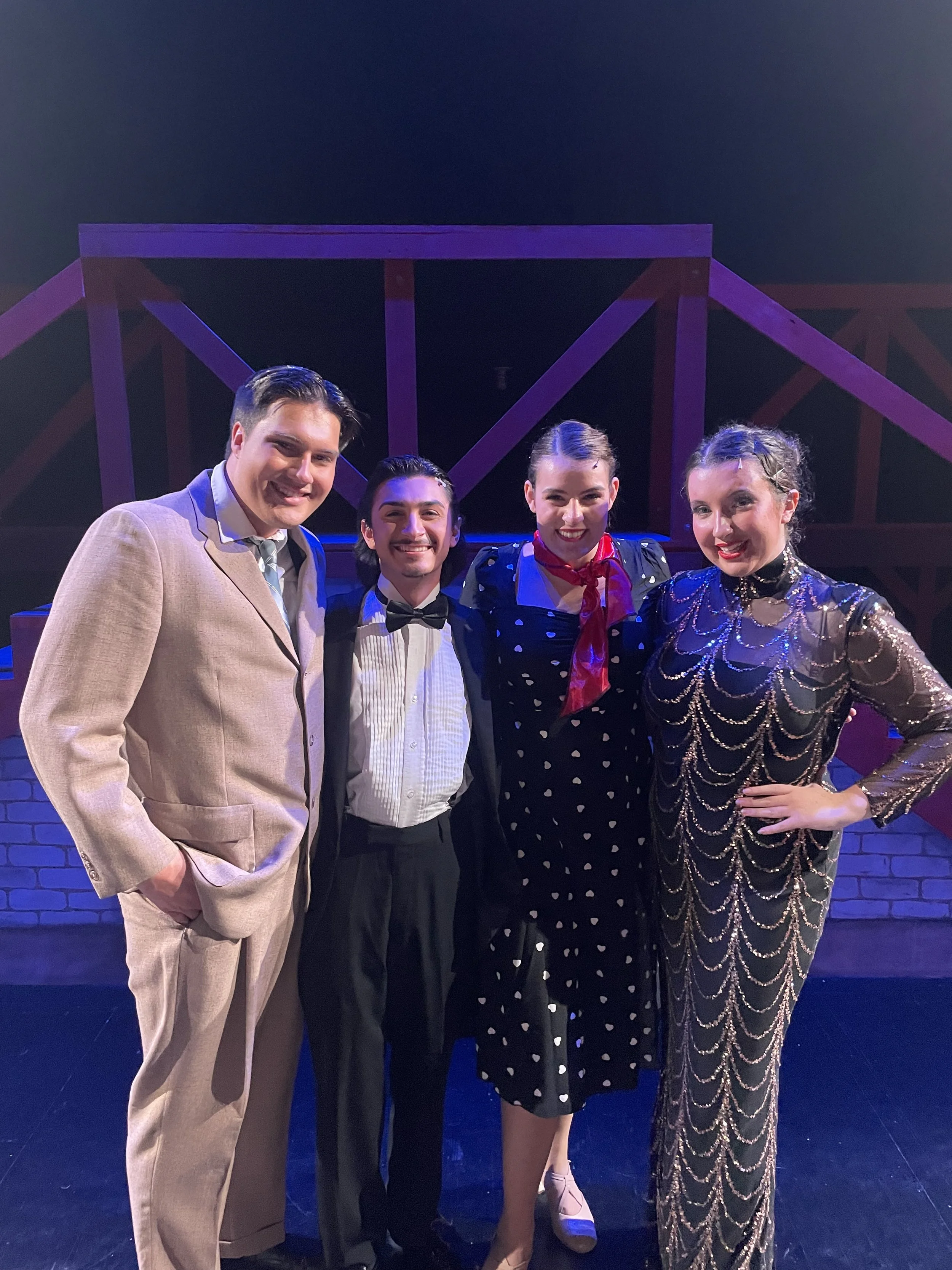 Four people dressed in vintage costumes standing together on stage, smiling at the camera with a set resembling a bridge in the background.