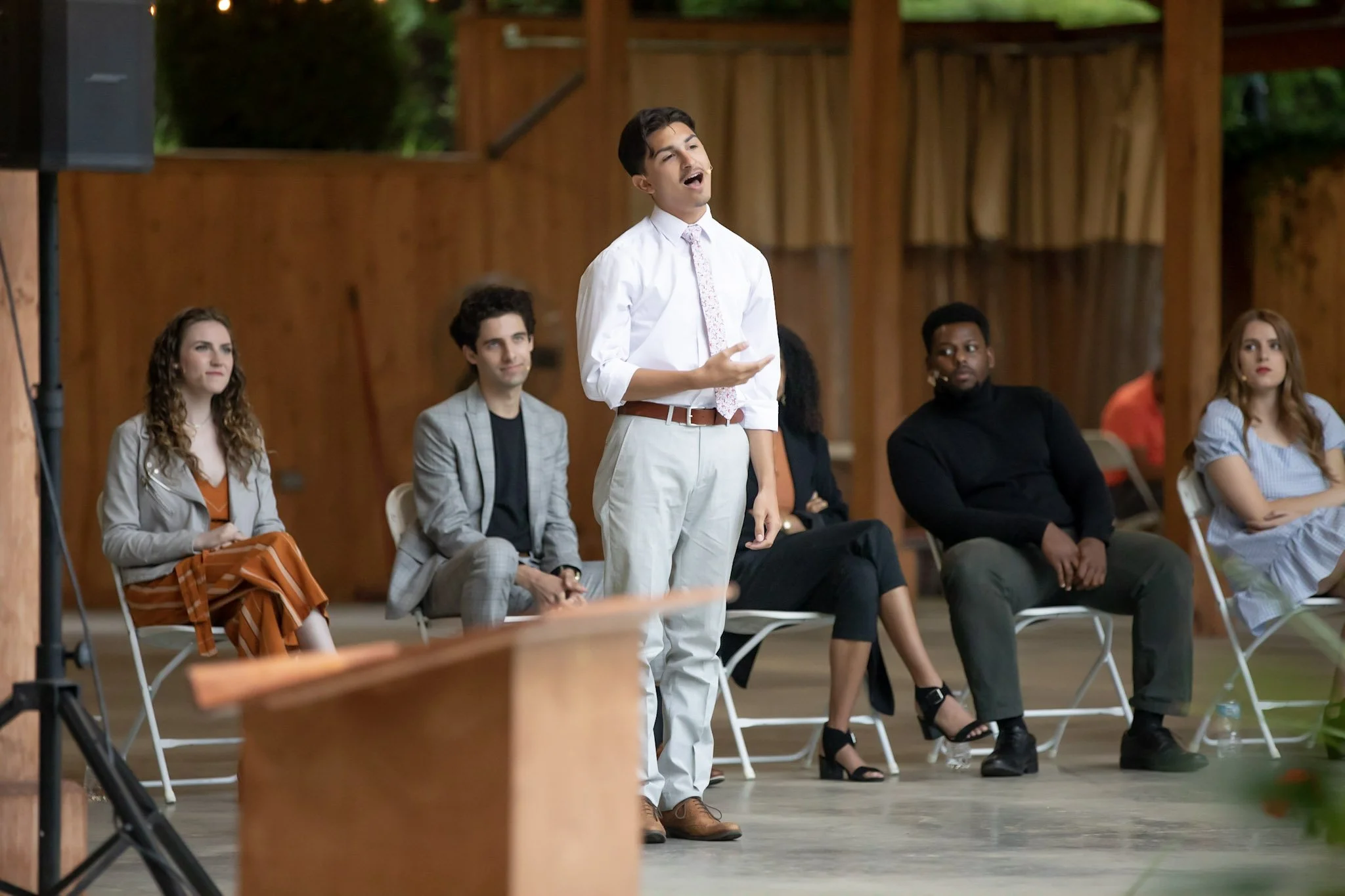 A young man wearing a white shirt, patterned tie, and beige pants is speaking or singing at an indoor event with a wooden background, seated audience members watch attentively.