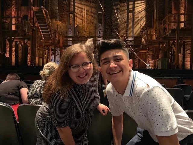 Two people smiling and leaning towards the camera in a theater or stage setting with wooden structures and theatrical lighting in the background.