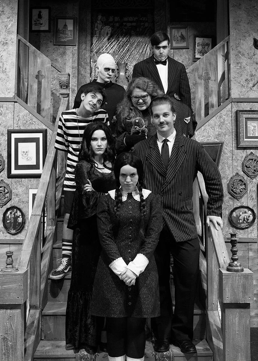 Group of seven people dressed in dark, vintage, or gothic-style clothing posing on a staircase. The setting appears to be decorated with framed pictures and vintage decor.