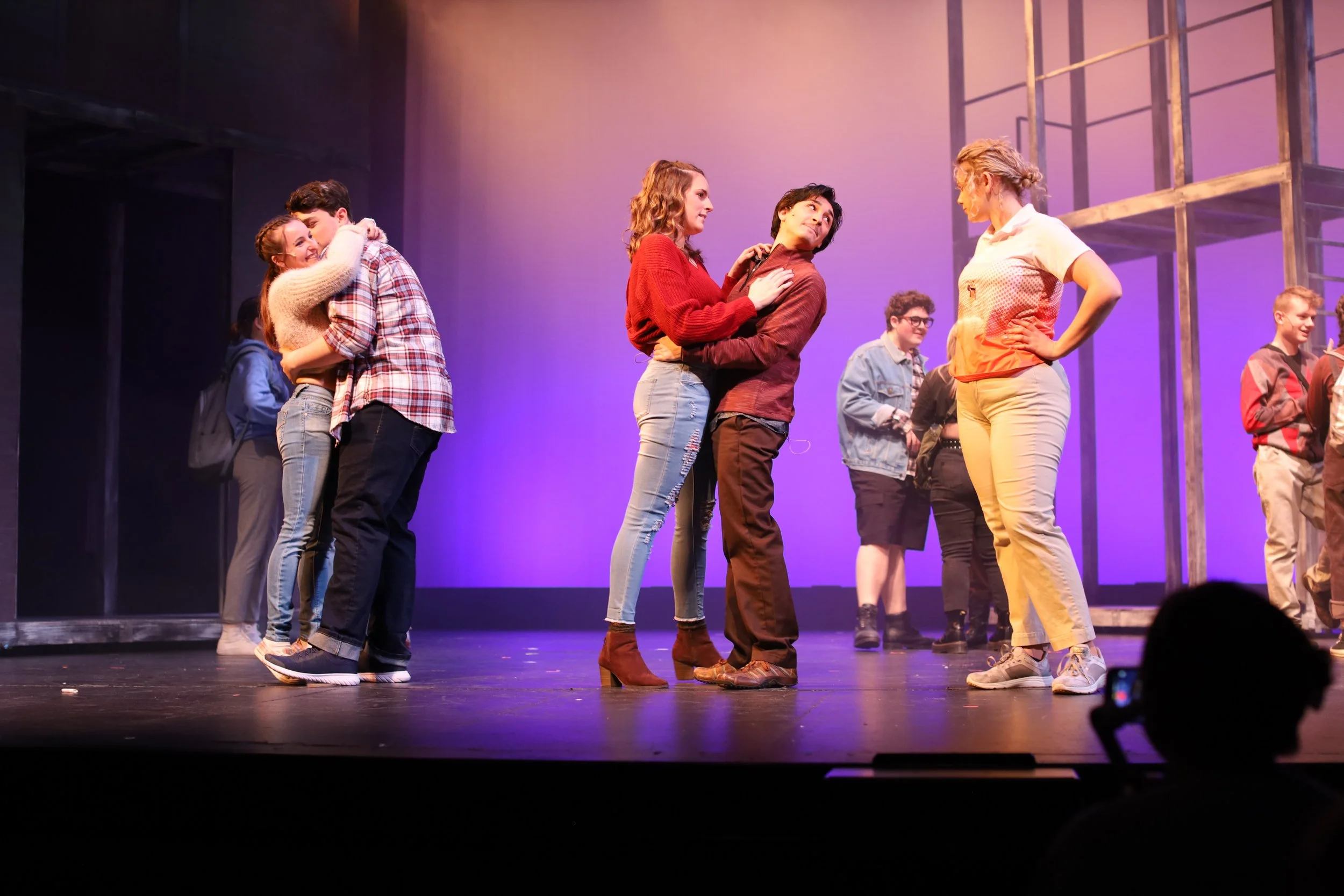 Theater stage with several young actors performing a play, with two couples in the foreground embracing or talking, and others in the background observing, under purple and yellow stage lighting.