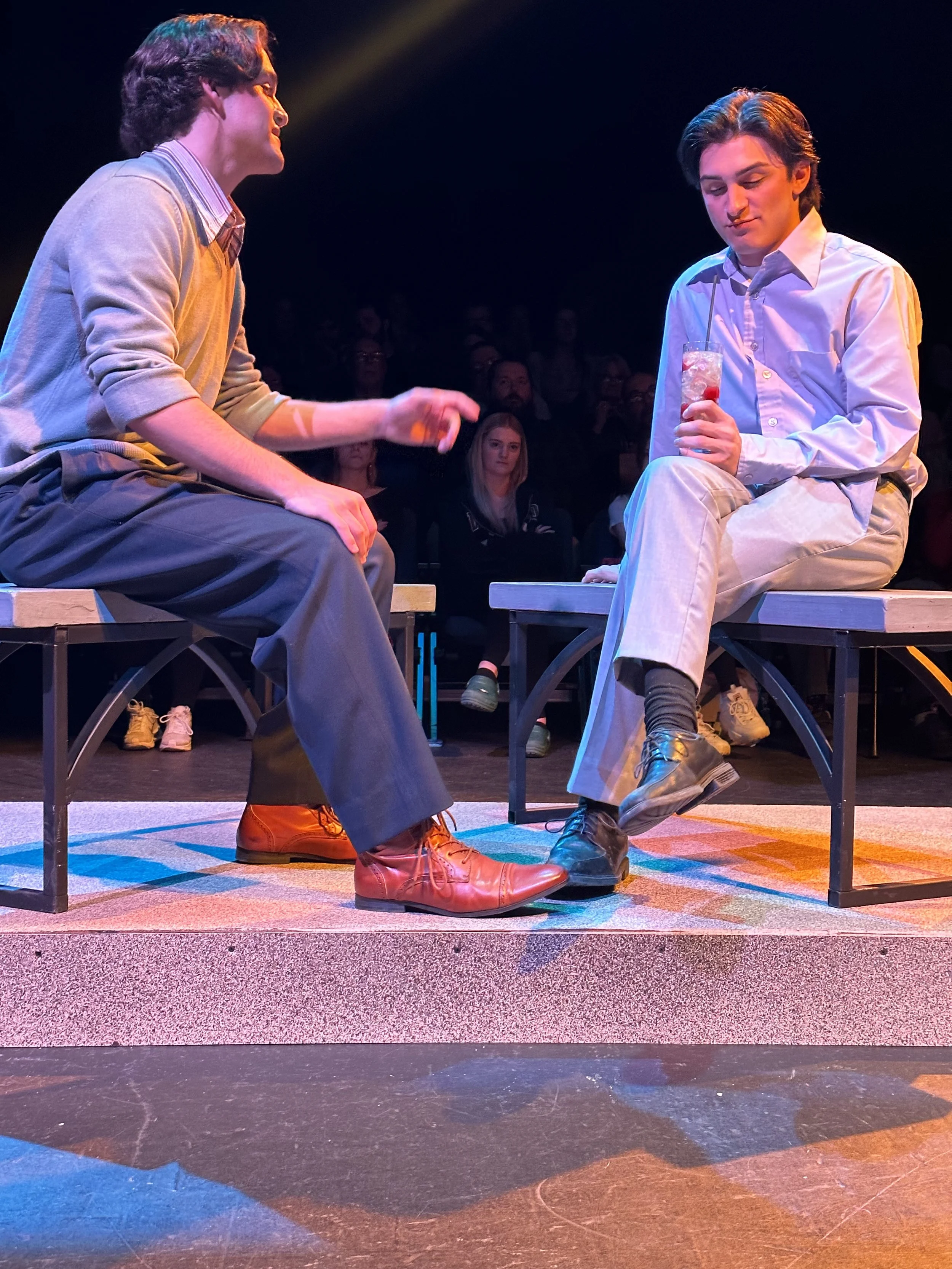 Two young men sitting on benches on stage, engaged in a conversation, with an audience in the background. One is holding a drink with a straw.