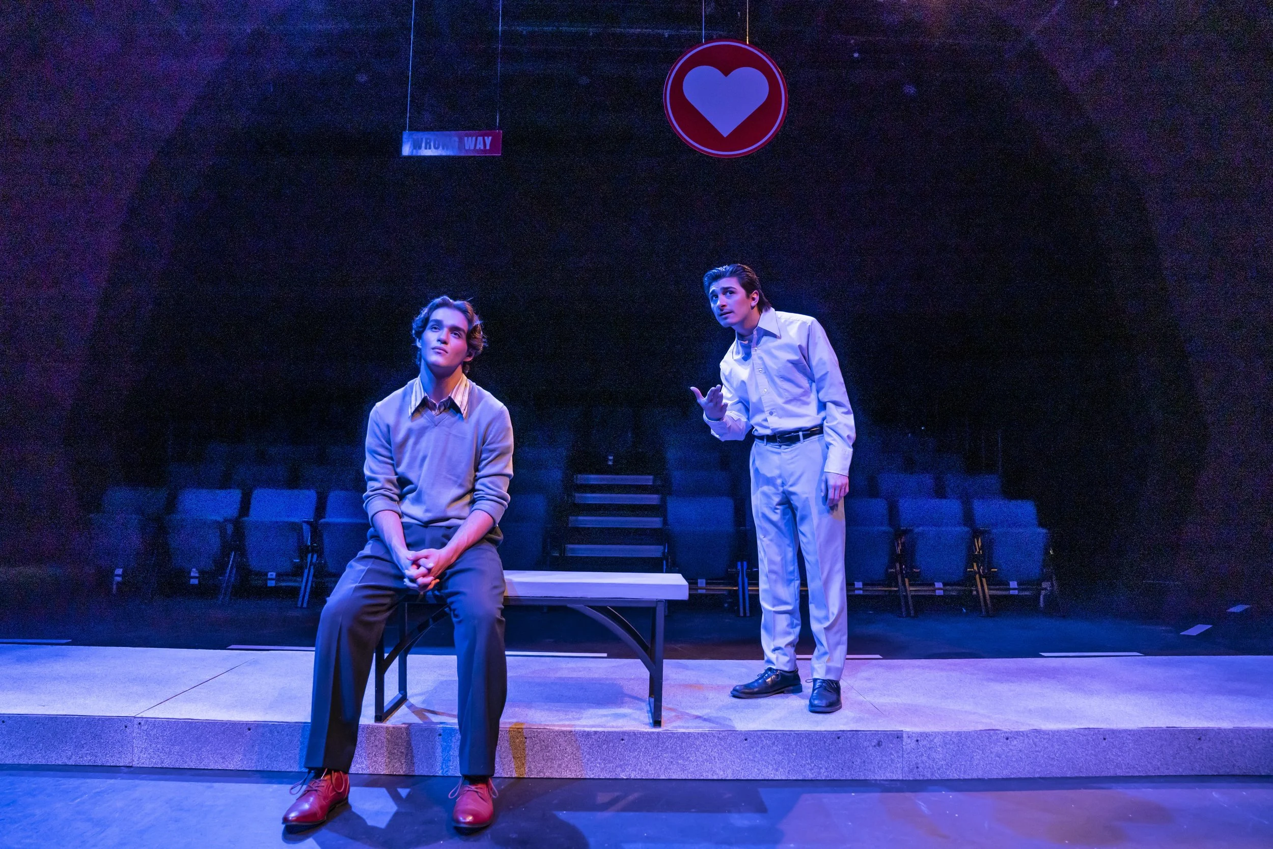 Two young actors on a stage with a dark background. One actor is sitting on a bench, looking contemplative, wearing a sweater and red shoes. The other actor is standing, gesturing with his hand, wearing a white shirt and white pants. There is a red a