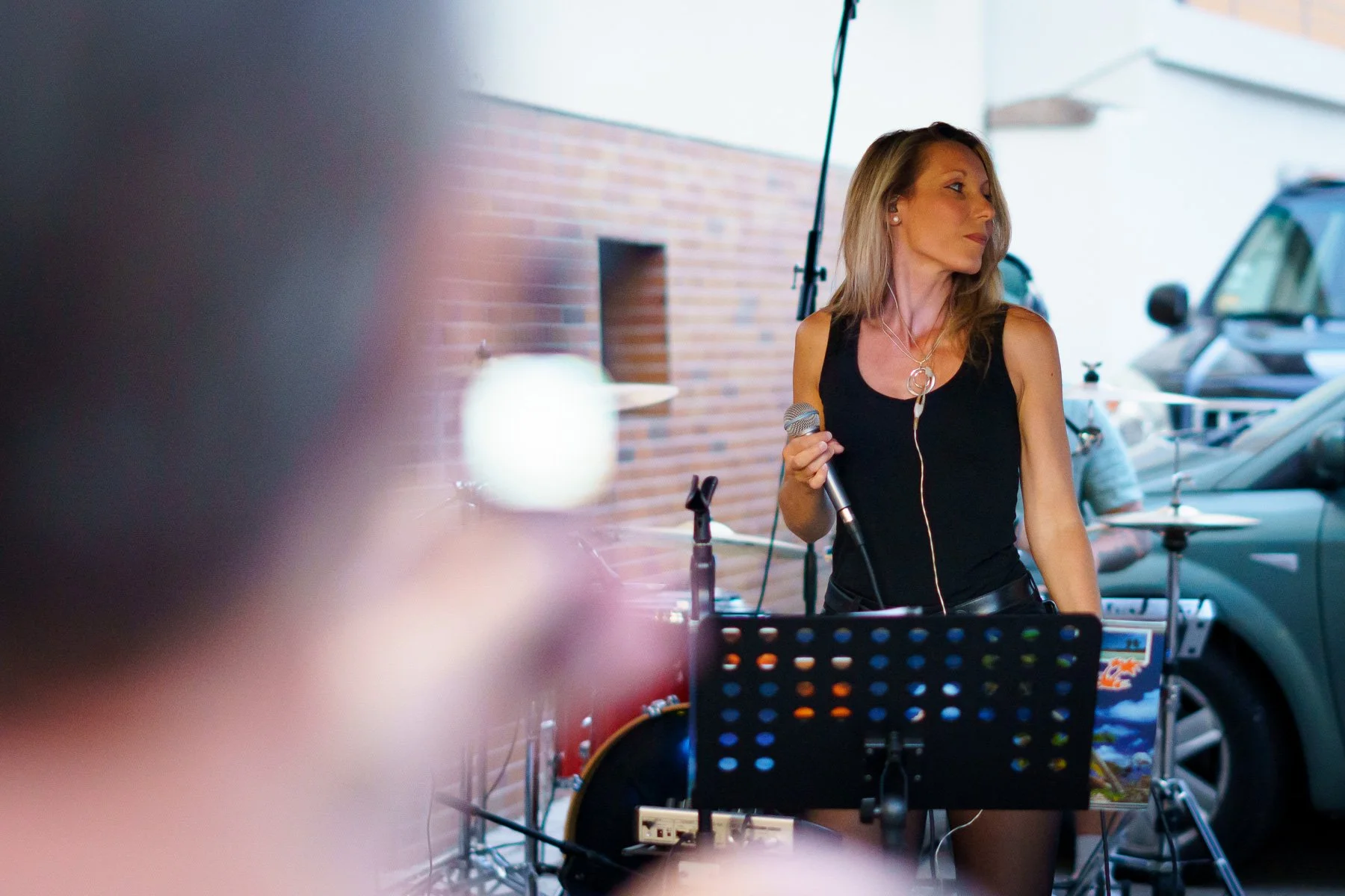 Une femme chante sur scène avec un microphone, devant un pupitre de musique, à côté d'une batterie. Elle est vêtue de noir et regarde sur le côté.