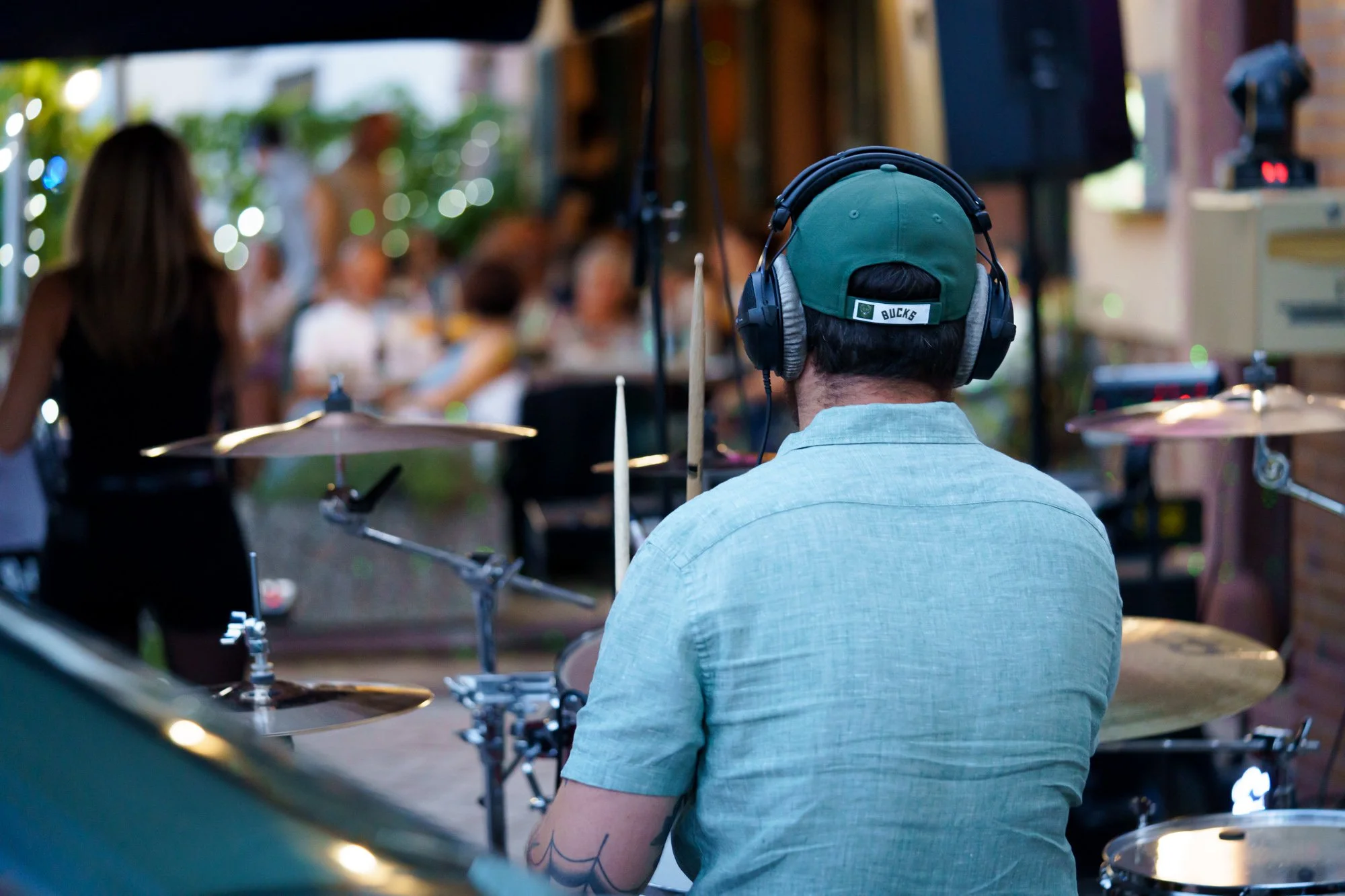 Un batteur portant un casque et une casquette verte joue de la batterie lors d'une performance en extérieur, entouré de spectateurs flous en arrière-plan.