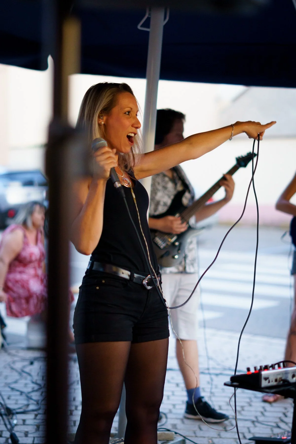Femme chantant sur scène avec un micro, doigt pointé, musicien jouant de la guitare en arrière-plan, équipement audio au sol.