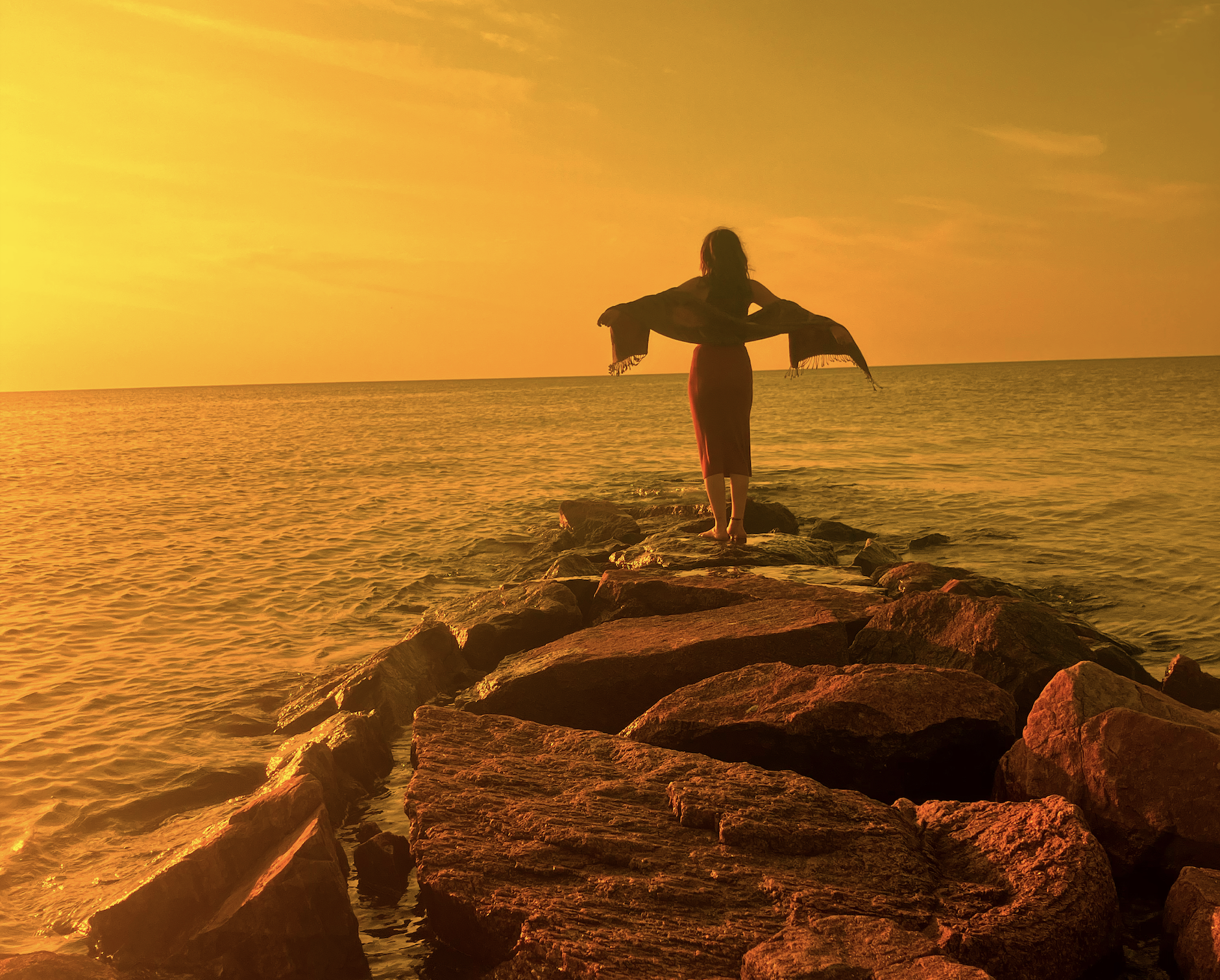 Person walking on rocks out to sea during sunset, arms outstretched, wearing a loose scarf and skirt.