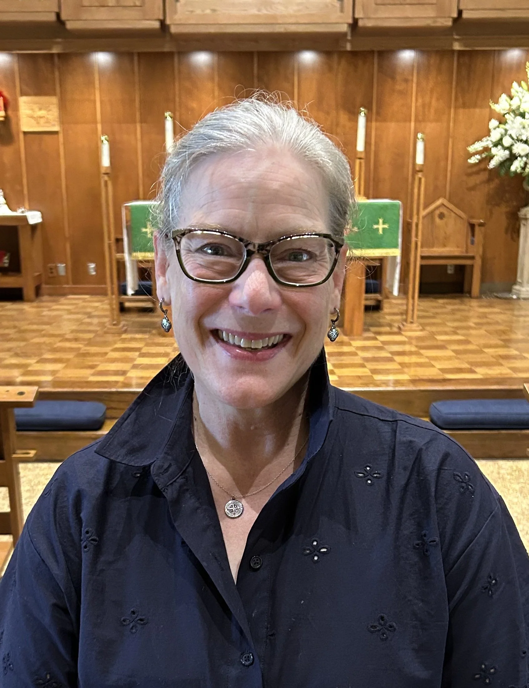 Smiling woman with glasses and earrings in a church interior with wooden panels, a green altar cloth, white flowers, and candles in the background.
