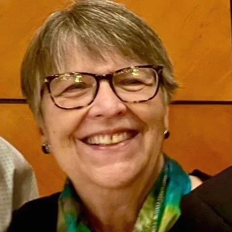 Close-up of a smiling middle-aged woman with short gray hair, glasses, and earrings, wearing a colorful scarf.