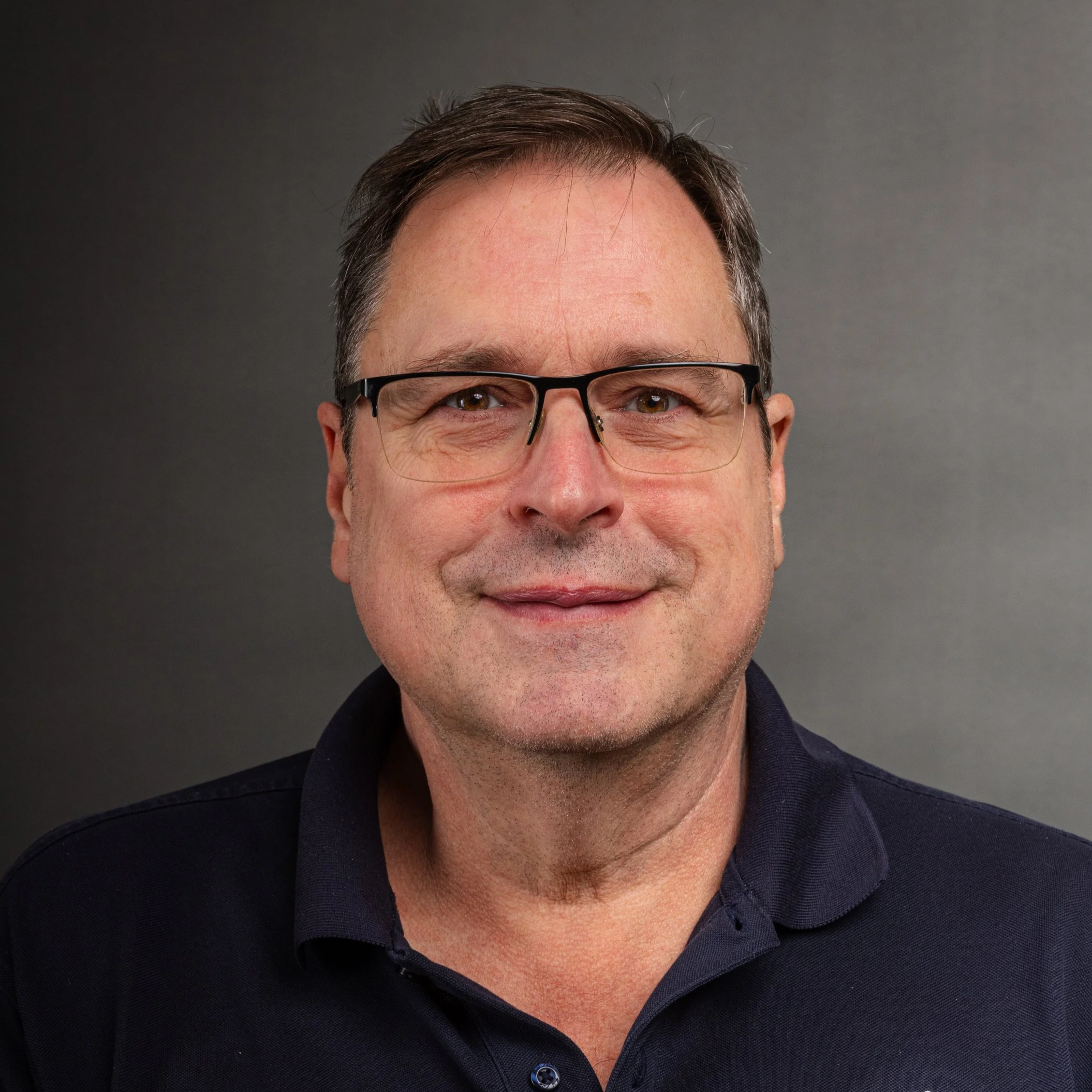 Close-up portrait of a middle-aged man with short dark hair, glasses, and a slight smile, wearing a dark collared shirt against a plain dark background.