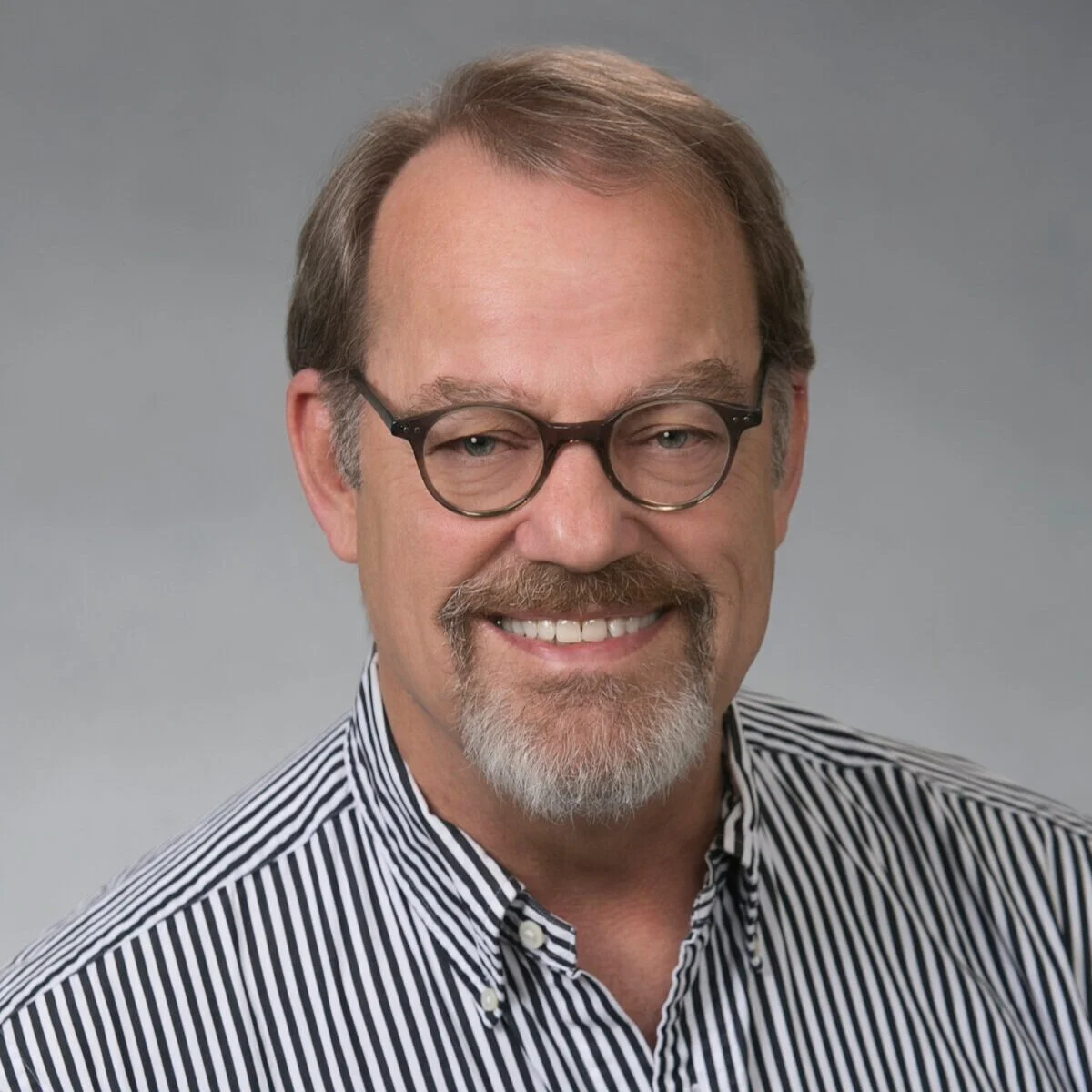 Portrait of a middle-aged man with light brown hair, gray beard, wearing glasses and a black-and-white striped shirt, smiling against a gray background.