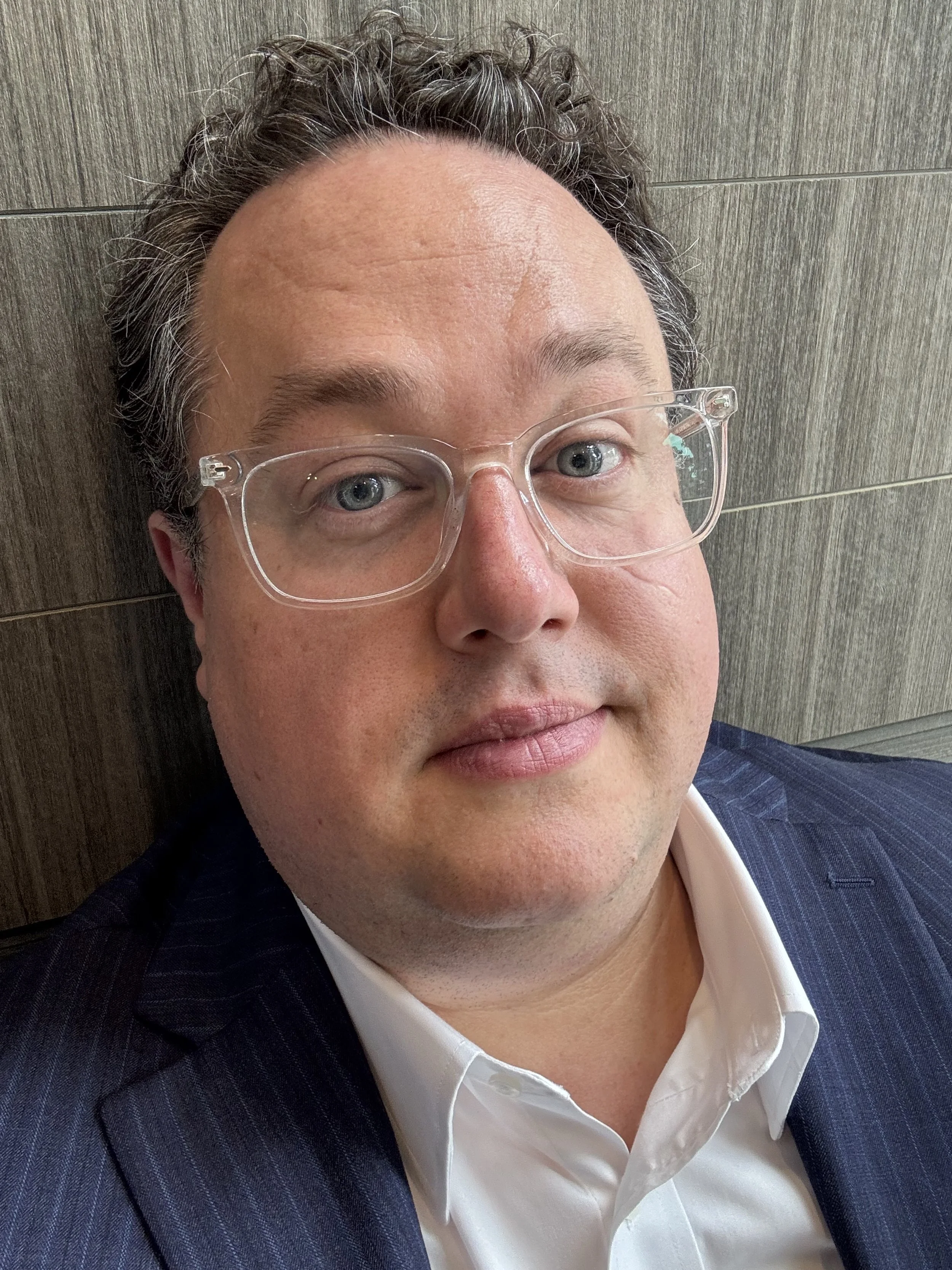 Close-up of a man with glasses, wearing a dark suit and white shirt, leaning against a wooden wall.