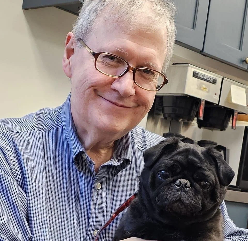 A smiling middle-aged man with glasses holding a black pug puppy indoors.