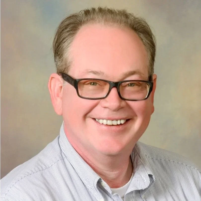 A smiling man with glasses and short, graying hair wearing a striped collared shirt against a neutral background.