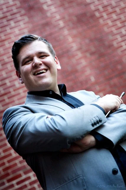 Smiling man in a grey suit with arms crossed, standing in front of a red brick wall.