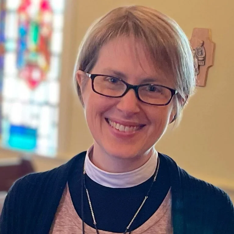 A smiling woman with short blonde hair, glasses, wearing a black cardigan over a pink shirt with a white clerical collar, in a church or similar setting with stained glass windows in the background.