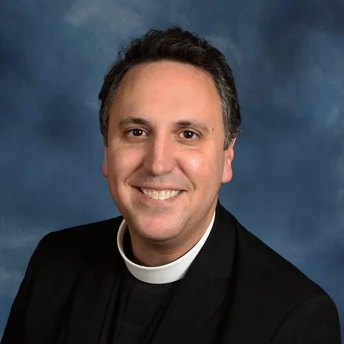 Portrait of a smiling man wearing clerical attire against a blue background.