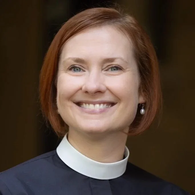 A woman with short red hair, blue eyes, and a wide smile, wearing a clerical collar and pearl earrings.