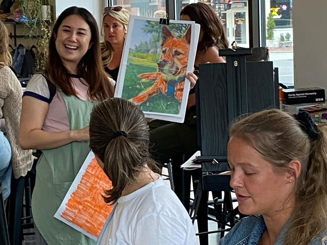 A woman in an art class holding a colorful drawing of a fox, smiling, with other students and easels in the background, creating a lively and creative atmosphere.