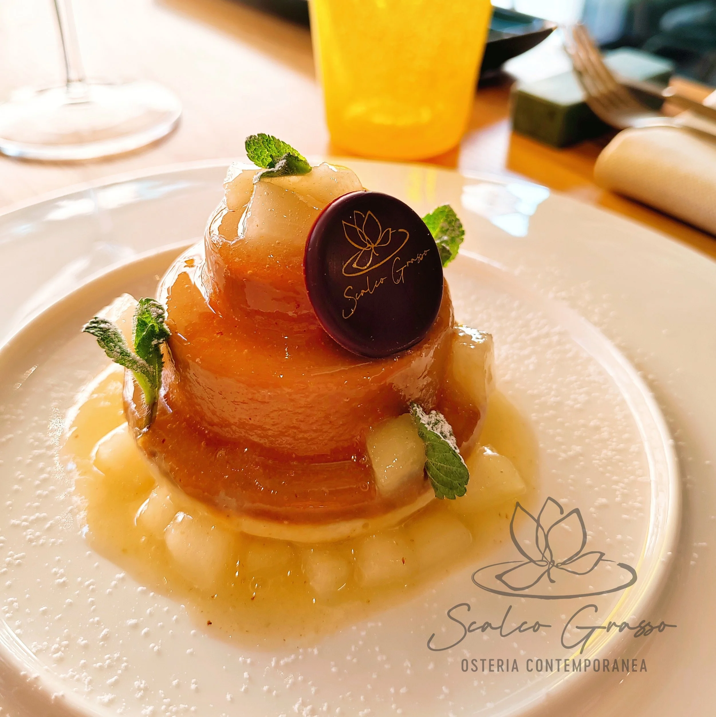 Dessert namelaka budino e foglie di menta, decorato con una medaglia di cioccolato con il logo 'Scalco Grasso' in un piatto bianco, con un bicchiere di bevanda gialla sullo sfondo.