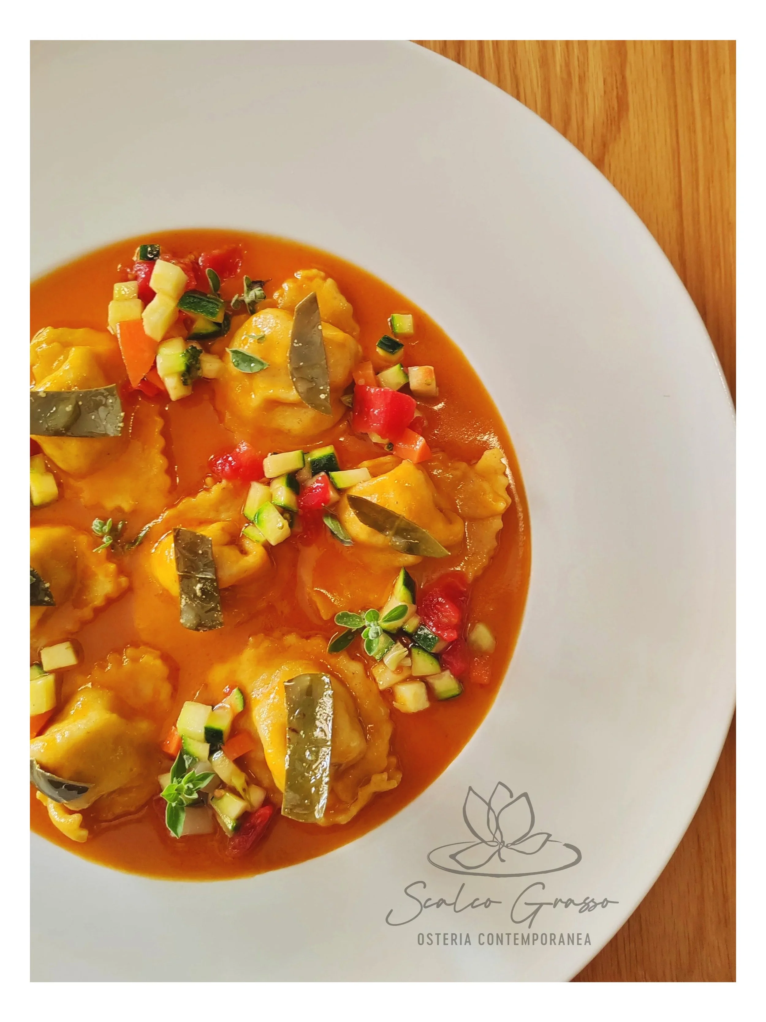 Piatto di ravioli ripieni con salsa di pomodoro, decorato con erbe fresche e cubetti di zucchine e peperoni rossi, in un piatto bianco con un logo di un fiore e la scritta 'Scalco Grasso Osteria Contemporanea'. Piatto estivo del nuovo menù estivo.