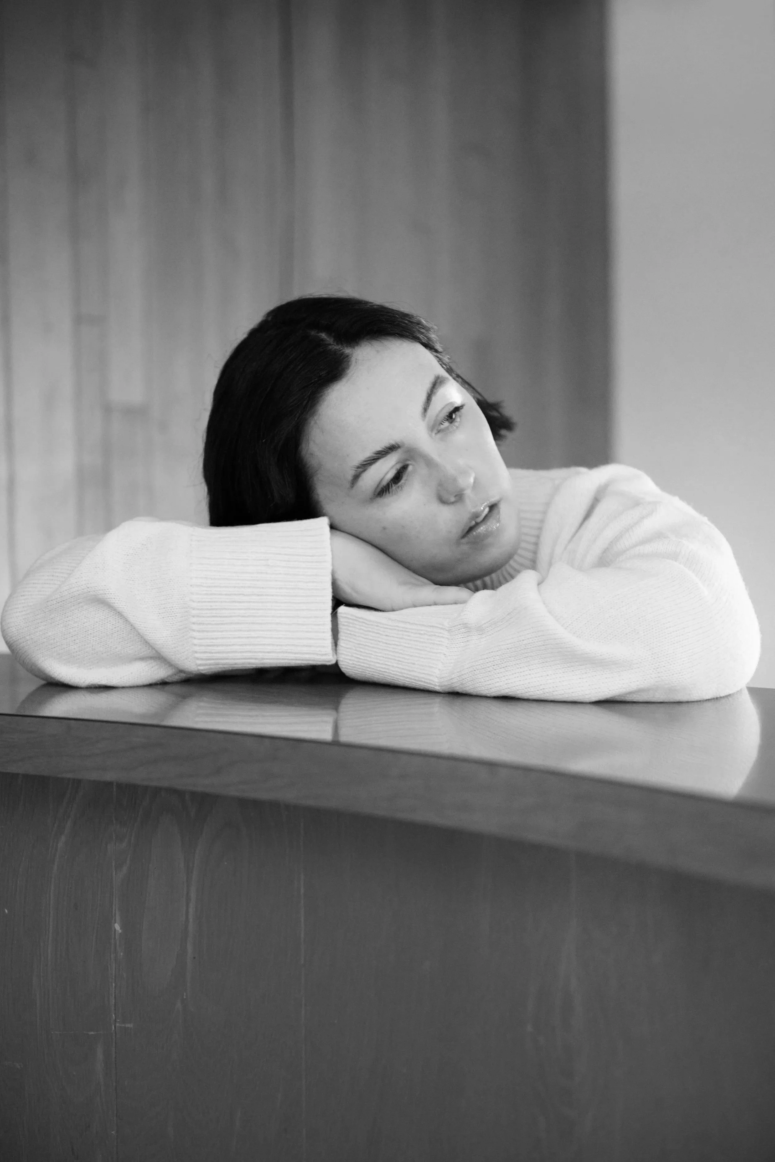 A young woman with dark hair resting her head on her arms on a table, looking contemplative in black and white.