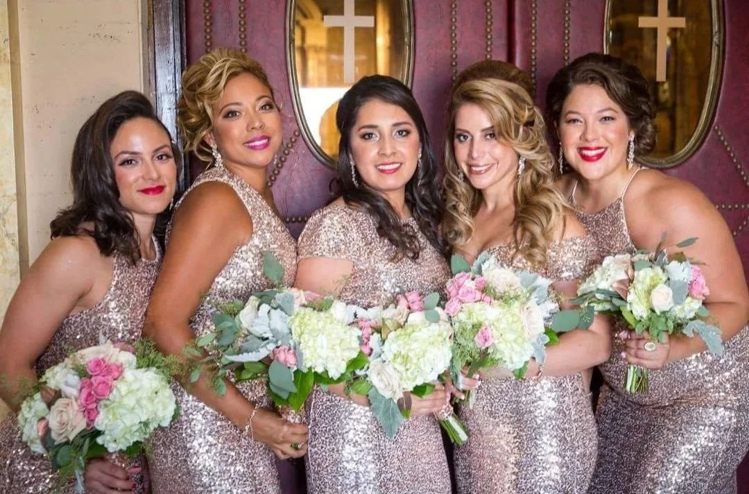 Six women in sparkling dresses holding bouquets of flowers, posing indoors in front of a decorated wall with mirrors.