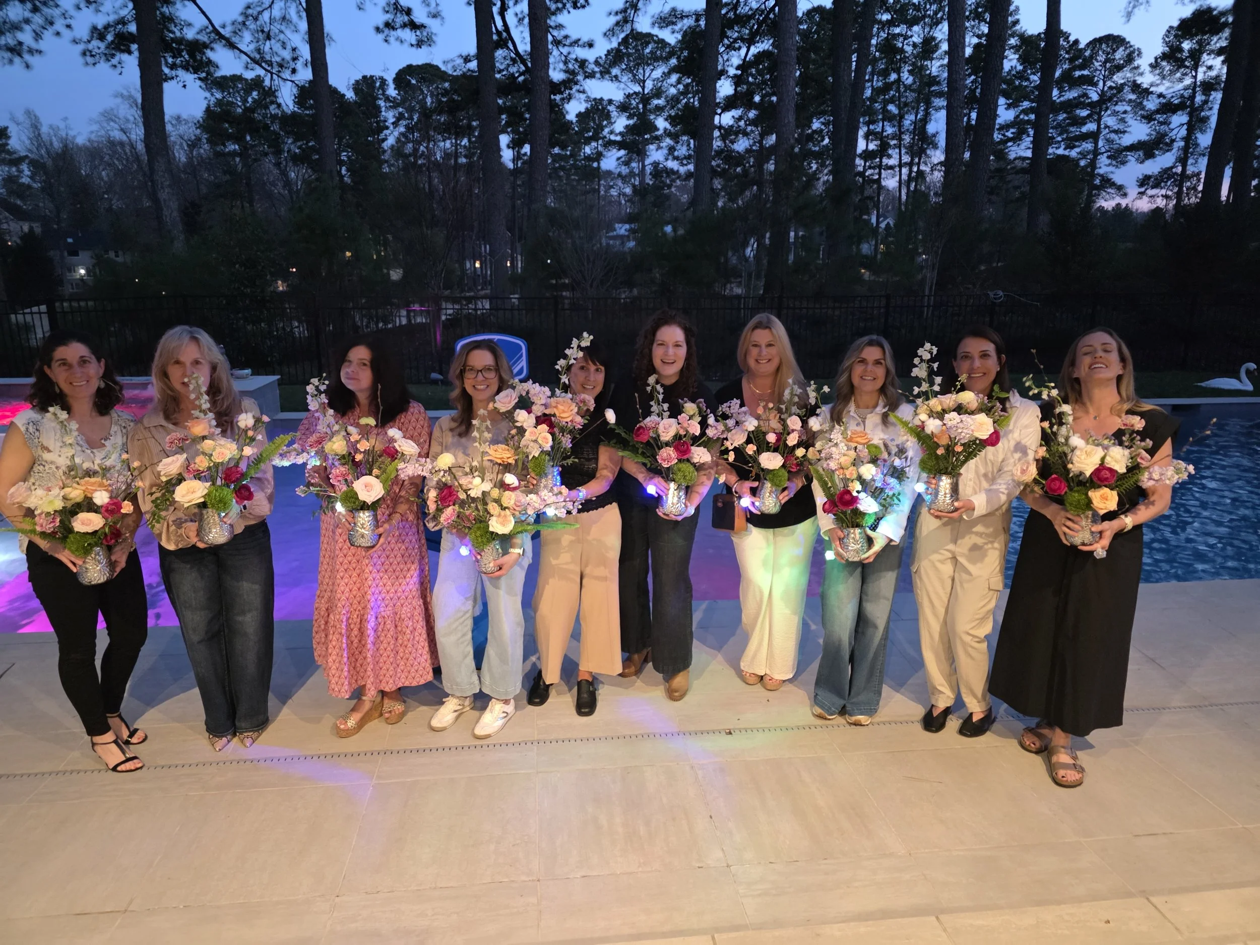 A group of eleven women standing side by side outdoors, holding flower arrangements in vases, with trees and a fence in the background during evening or dusk.