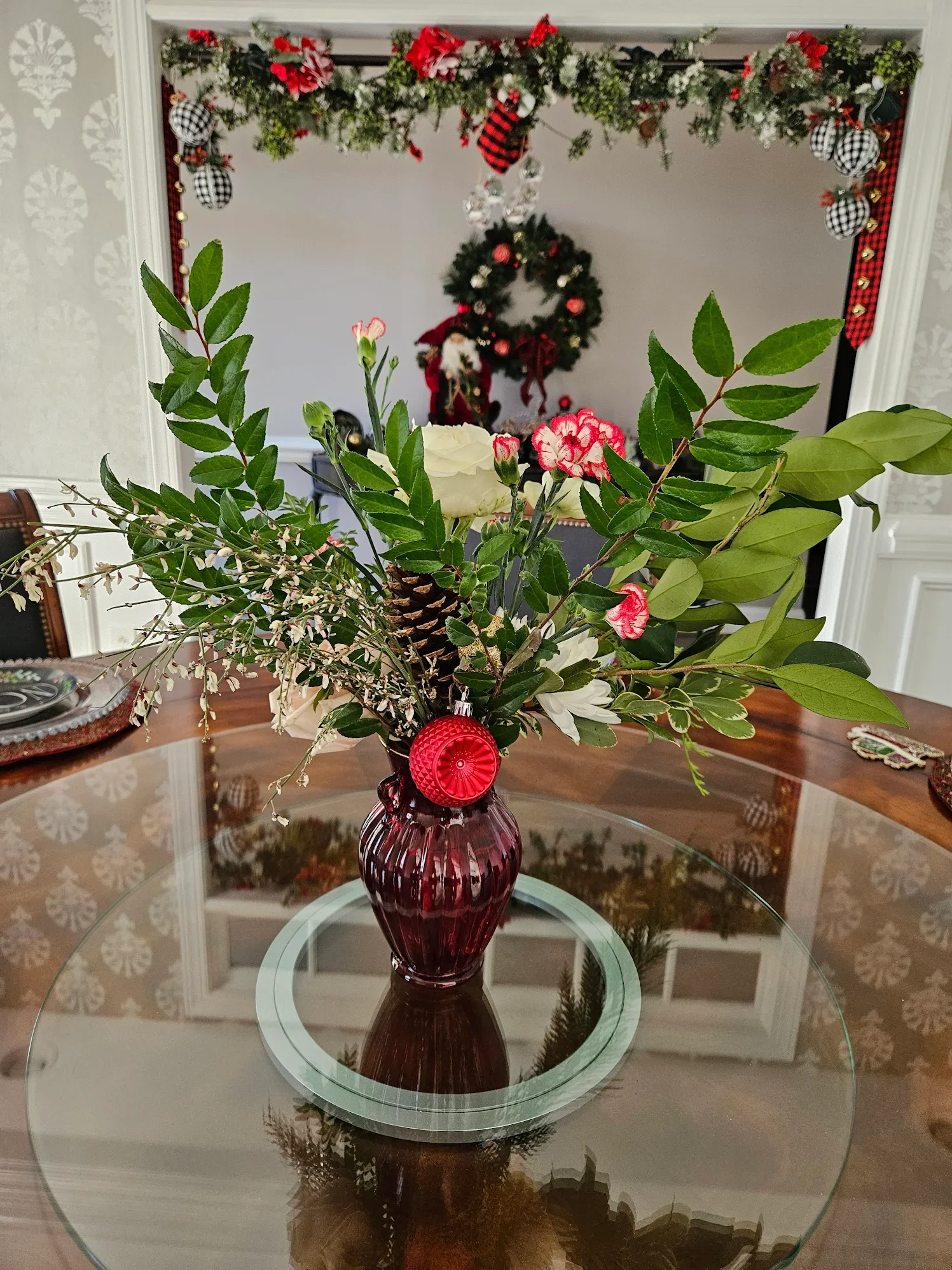 A glass vase with red color and ribbed design holding an arrangement of green leaves, white flowers, red and white carnations, and small white blossoms, placed on a glass table with a decorative mirror reflection.