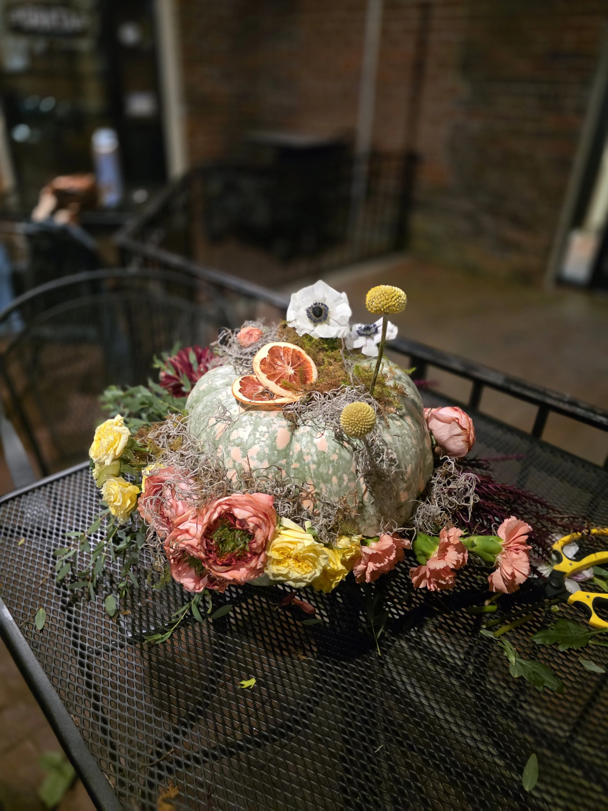 A floral arrangement on top of a pumpkin, including roses, anemones, dried orange slices, and other dried flowers and foliage, on a black metal outdoor table.