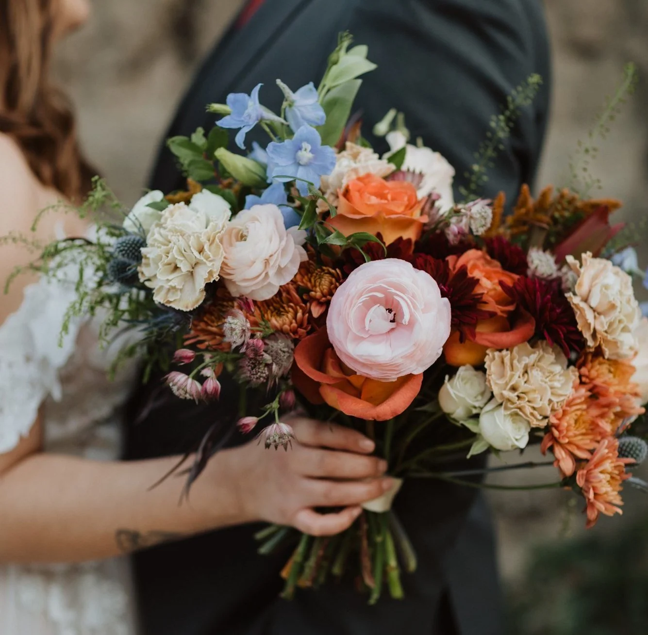 A person holding a large bouquet of mixed colorful flowers, including pink, peach, white, and blue blossoms, with a blurred background.