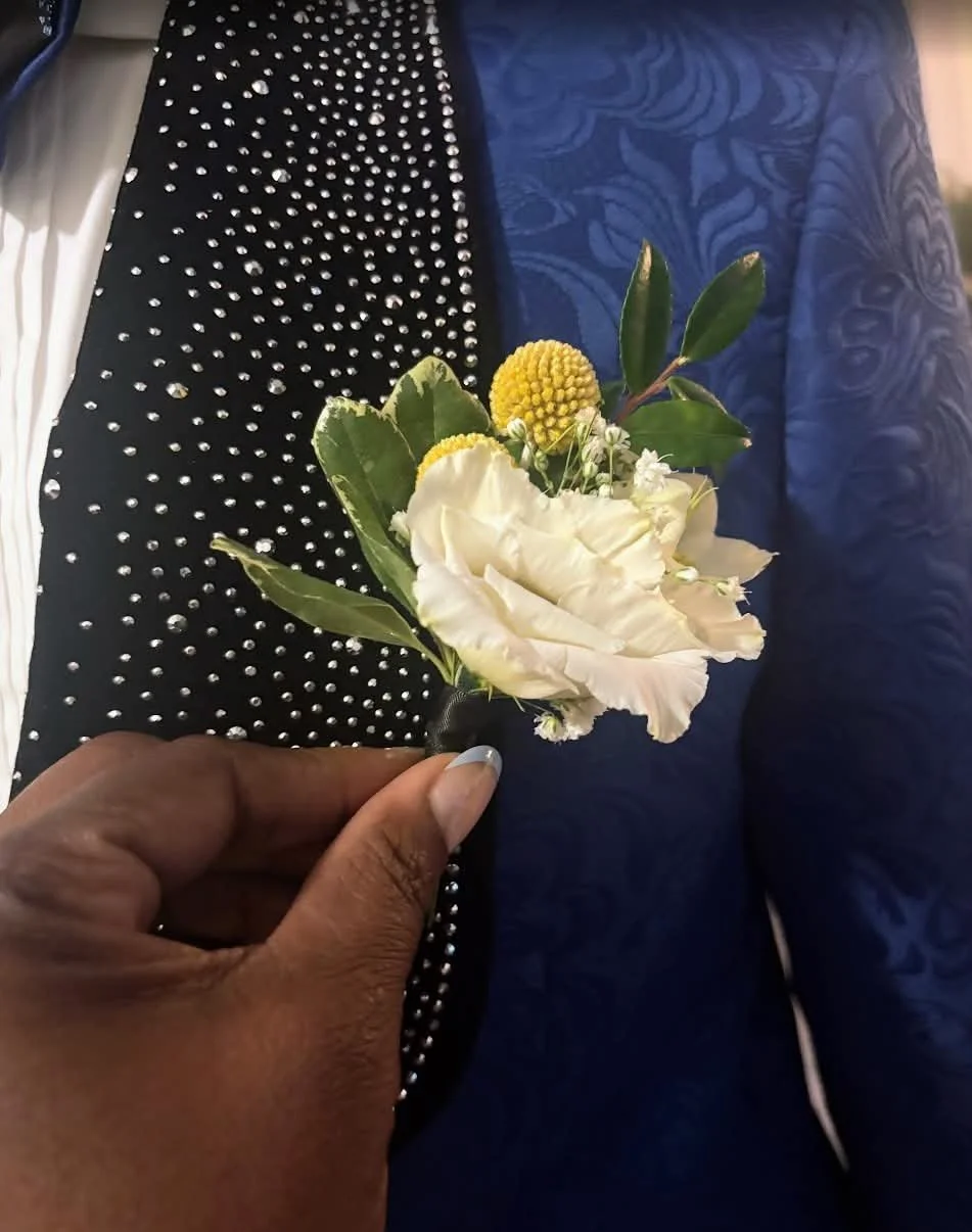 Person holding a small bouquet of white and yellow flowers with green leaves, against a background of black fabric with silver embellishments and a blue patterned fabric.