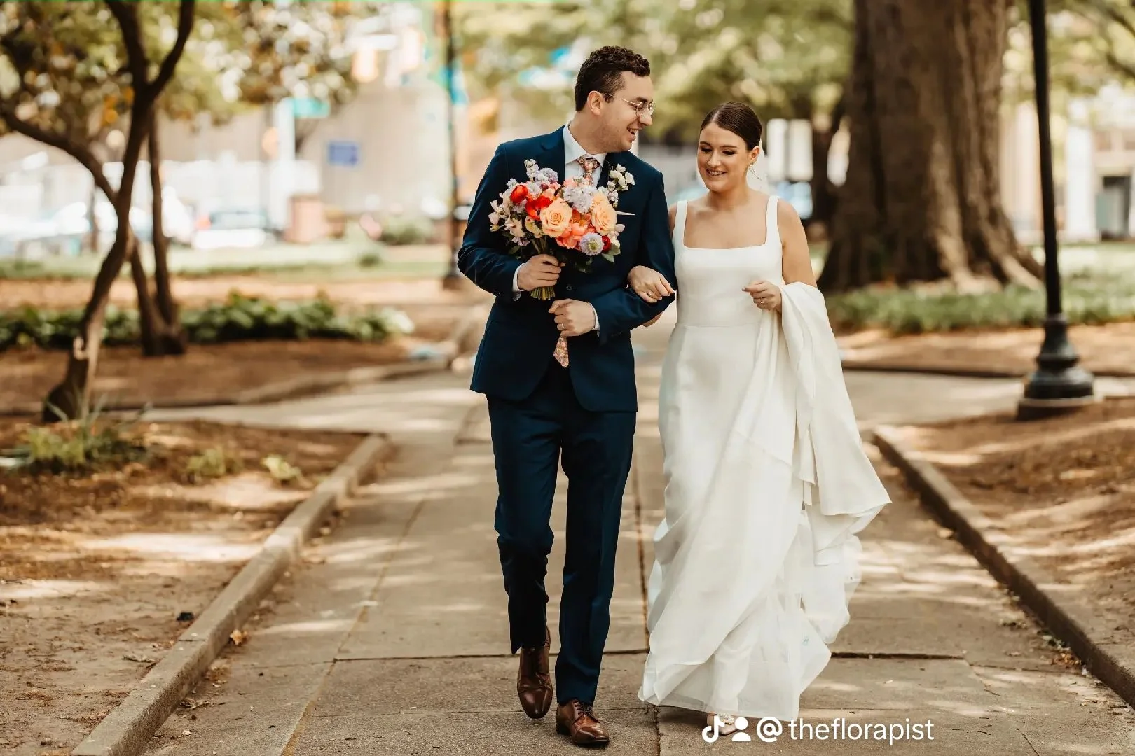 A couple in wedding attire walking arm-in-arm in a park with trees and a pathway.