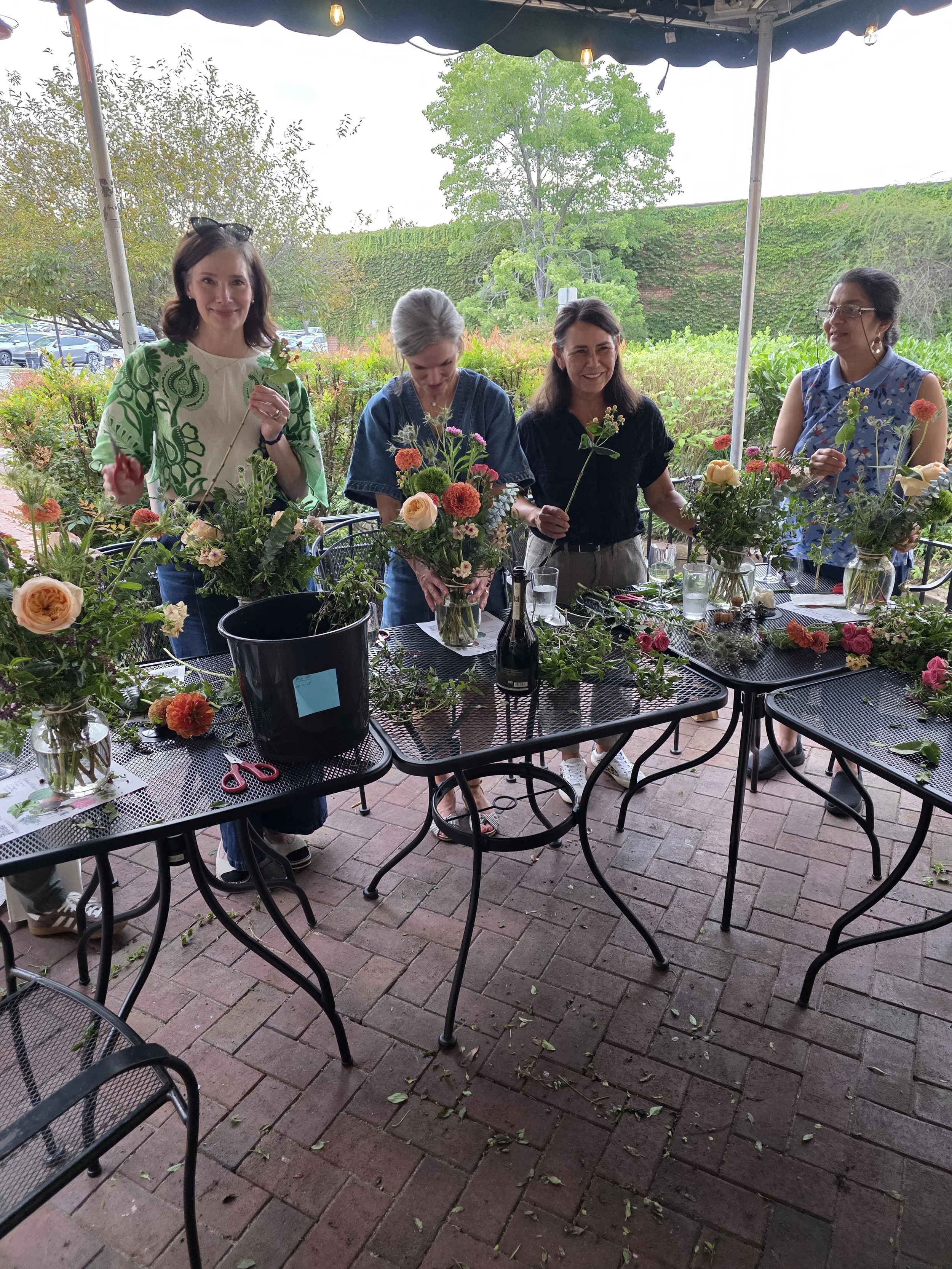 Four women standing outdoors under a canopy, surrounded by flower arrangements on black tables with glassware and scissors, in a garden setting with trees and a brick patio.