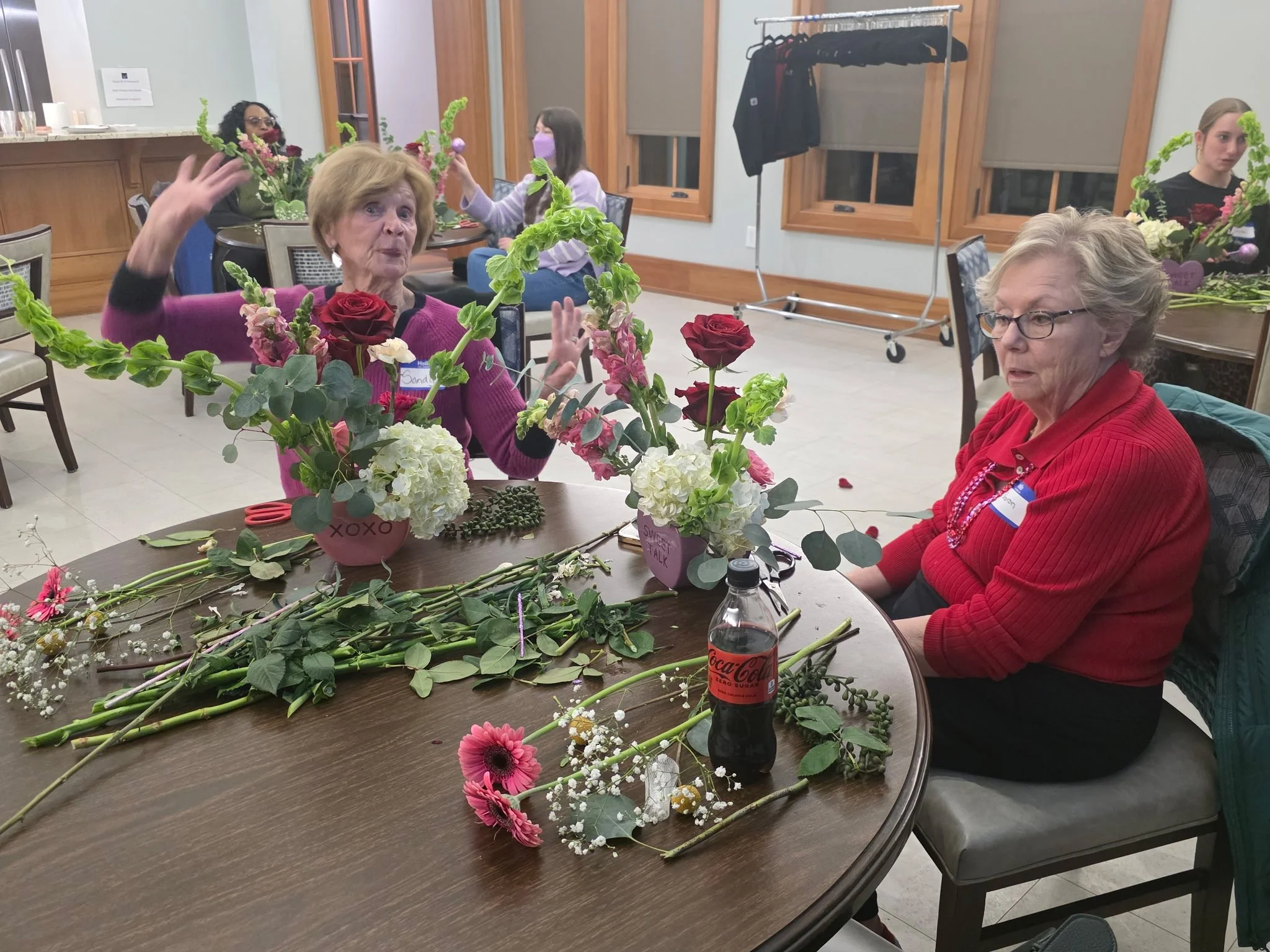 Women decorating a table with flowers, including roses, green foliage, and various other blooms, in a room with wood-paneled windows and a coat rack.