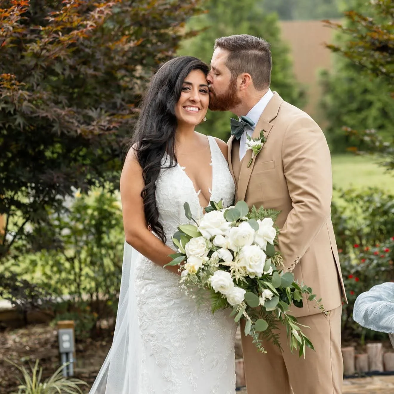 A newlywed couple standing outdoors, the groom kissing the bride's forehead, the bride holding a large bouquet of white roses and greenery, both smiling, surrounded by lush greenery and trees.